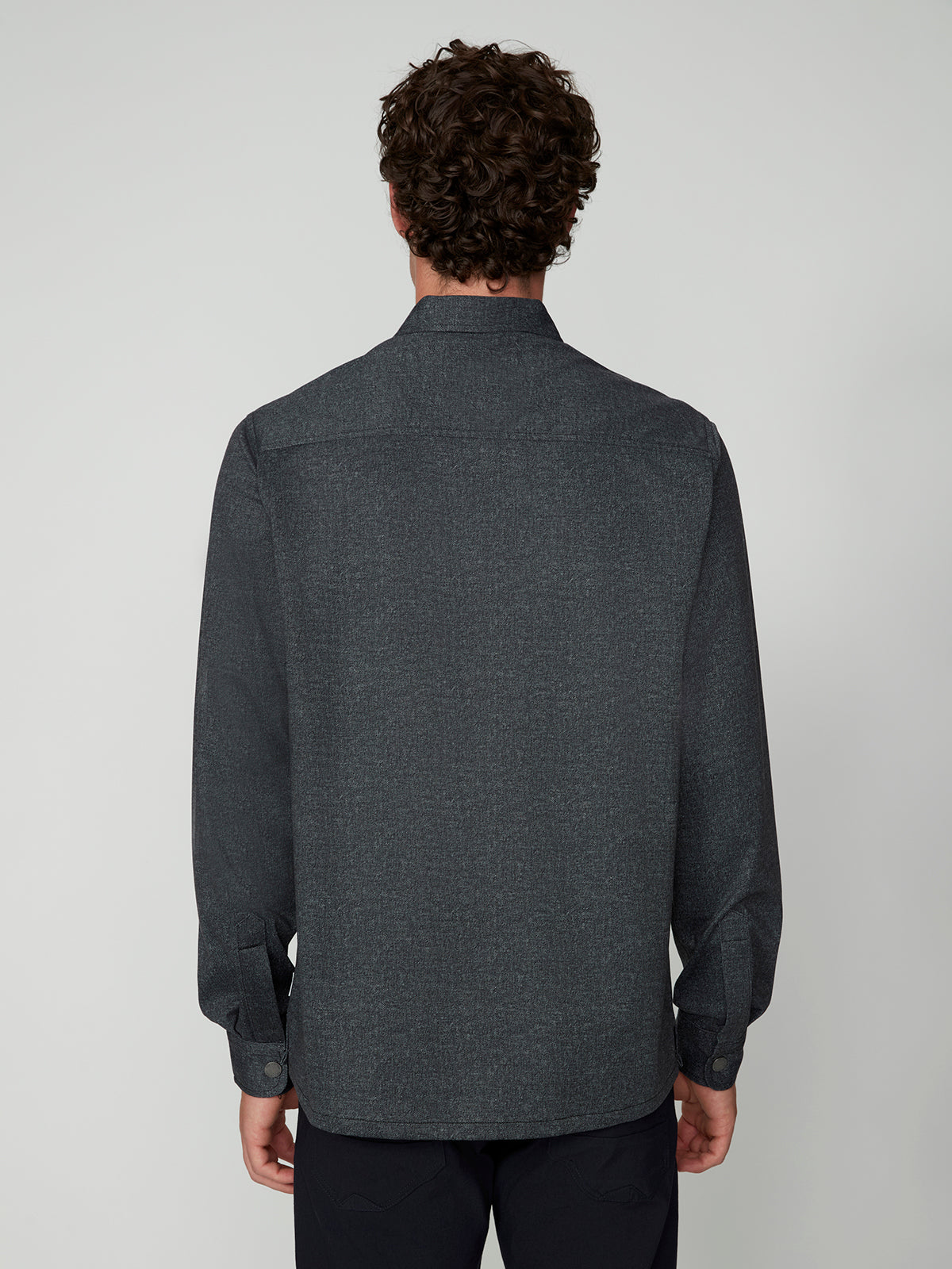 A man with curly brown hair is shown from behind, wearing a PROJEK RAW MENS LONG SLEEVES KNIT SHIRT and dark pants, standing against a plain light gray background.