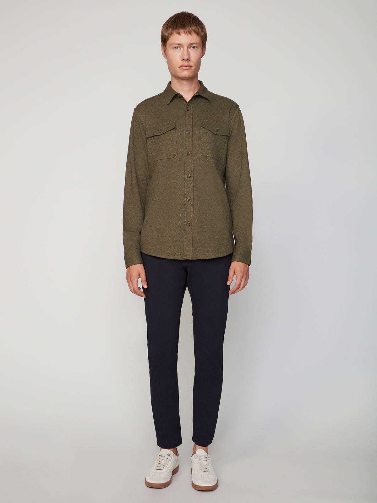 A man faces forward against a plain background, wearing a PROJEK RAW MENS LONG SLEEVES KNIT FLANNEL SHIRT in olive green, paired with dark navy pants and white sneakers.