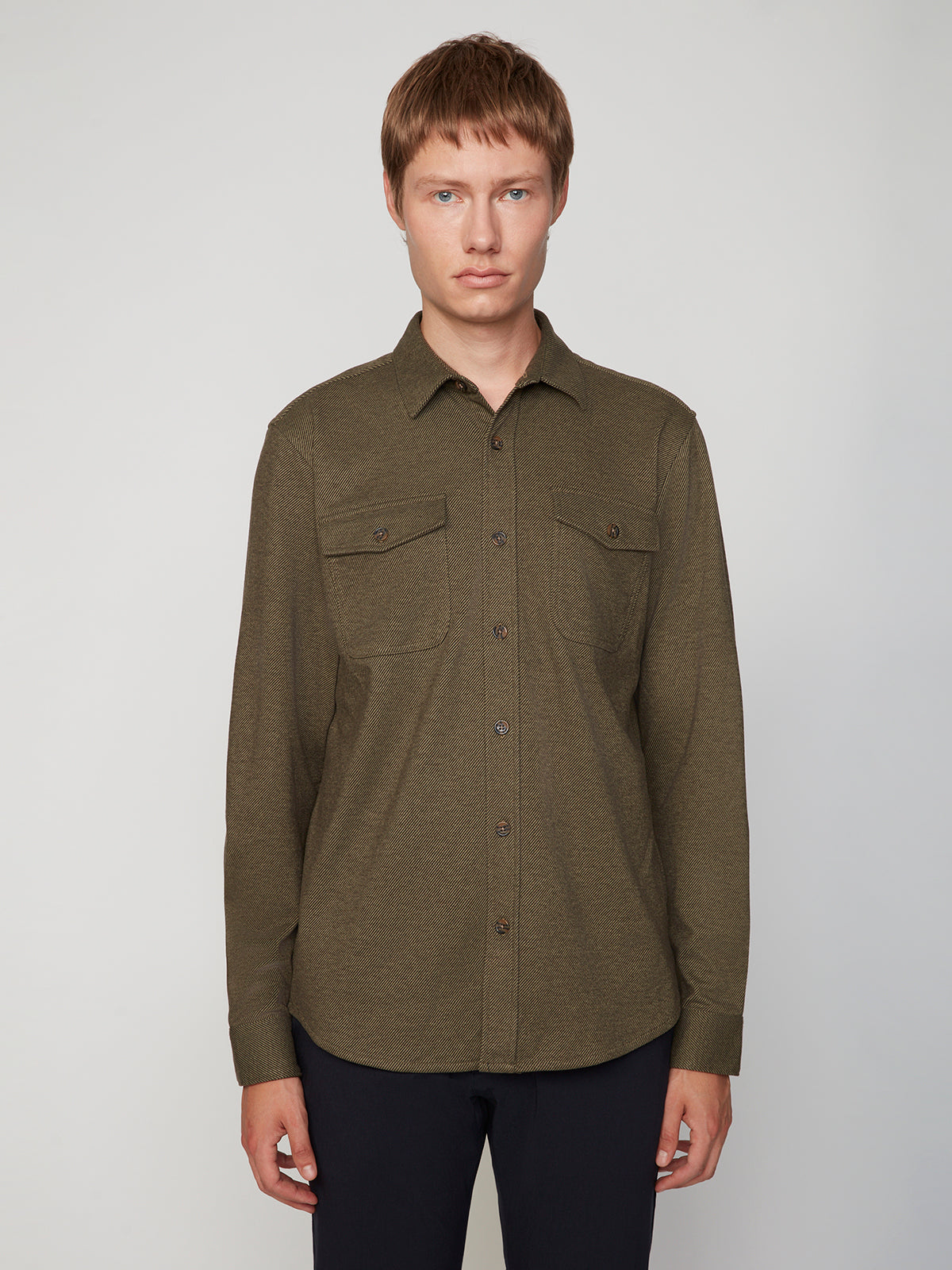 A young man with short light brown hair wears the PROJEK RAW MENS LONG SLEEVES KNIT FLANNEL SHIRT in olive green with two chest pockets and dark pants, standing against a plain light gray background, looking at the camera.