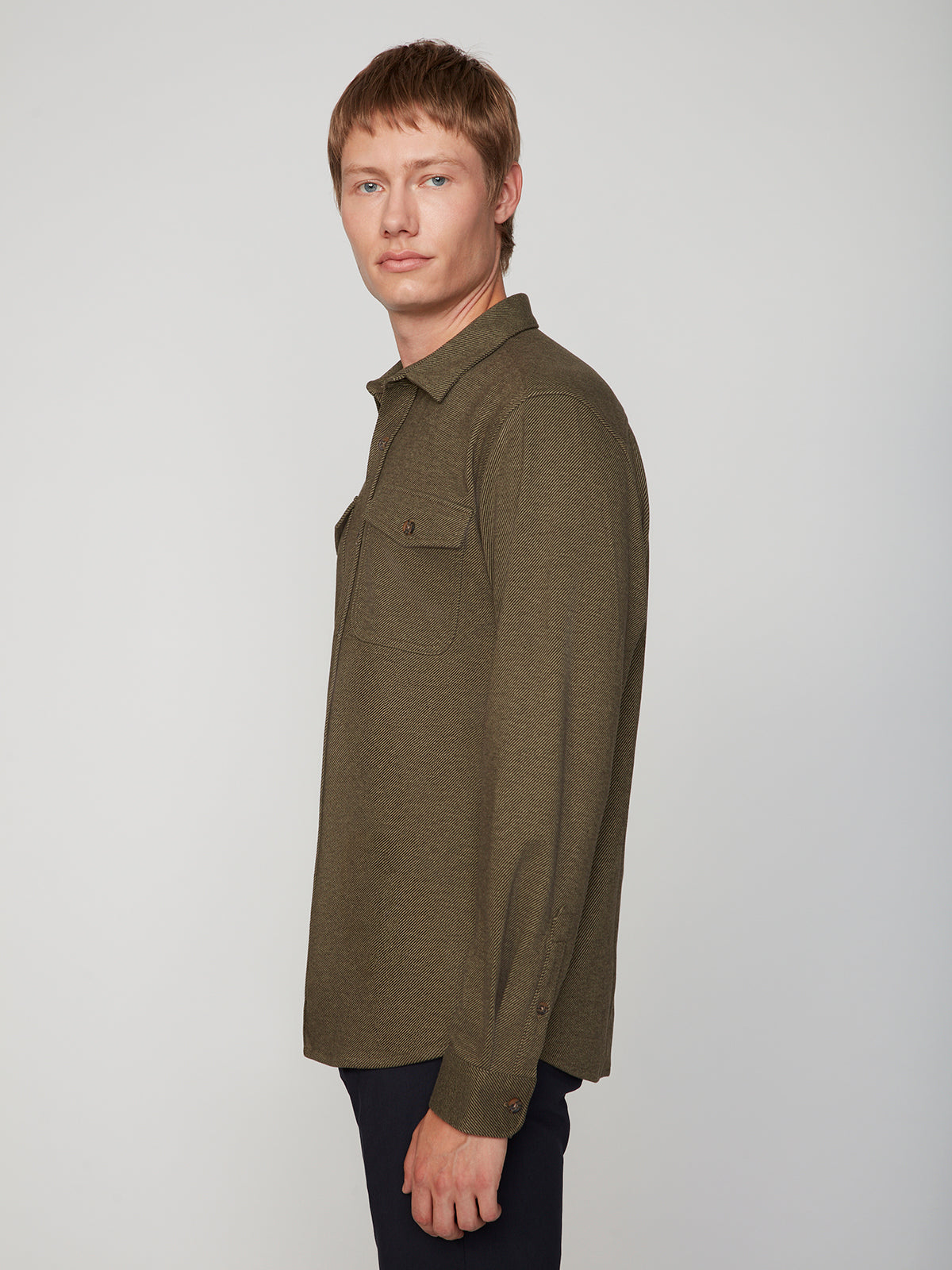 A young man with short light brown hair models the PROJEK RAW MENS LONG SLEEVES KNIT FLANNEL SHIRT in olive green, paired with dark pants and posed sideways against a plain light gray background, looking at the camera.