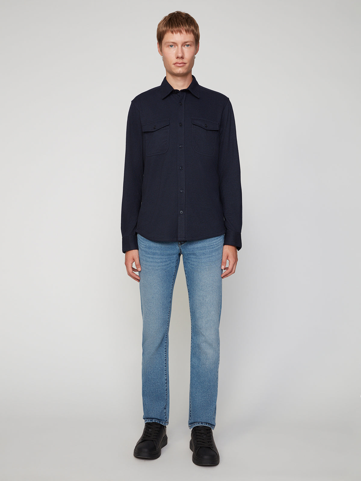 A person with short light brown hair stands facing forward, wearing a PROJEK RAW MENS LONG SLEEVES KNIT FLANNEL SHIRT, light blue jeans, and black shoes against a plain light background.