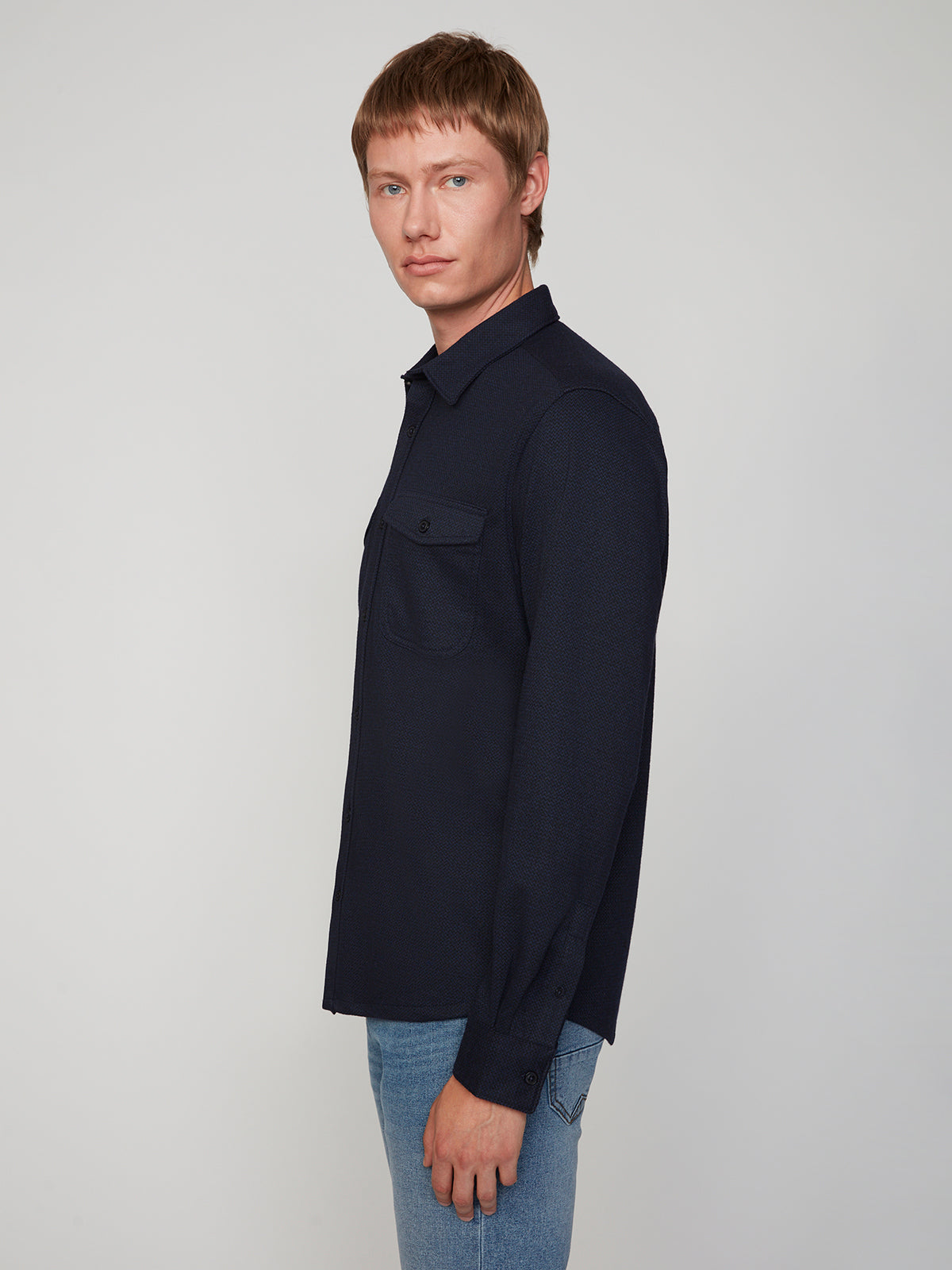 A young man with short light brown hair models the PROJEK RAW MENS LONG SLEEVES KNIT FLANNEL SHIRT and blue jeans, standing sideways and looking at the camera against a plain light background.
