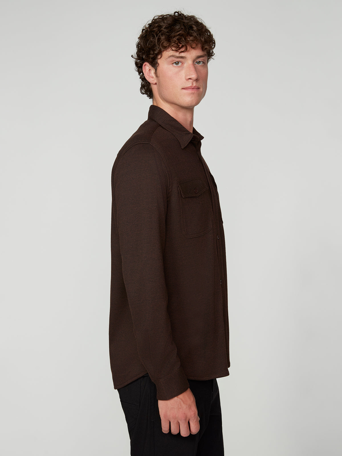 A young man with curly brown hair stands in profile against a plain background, wearing the PROJEK RAW MENS LONG SLEEVES KNIT FLANNEL SHIRT and black pants.