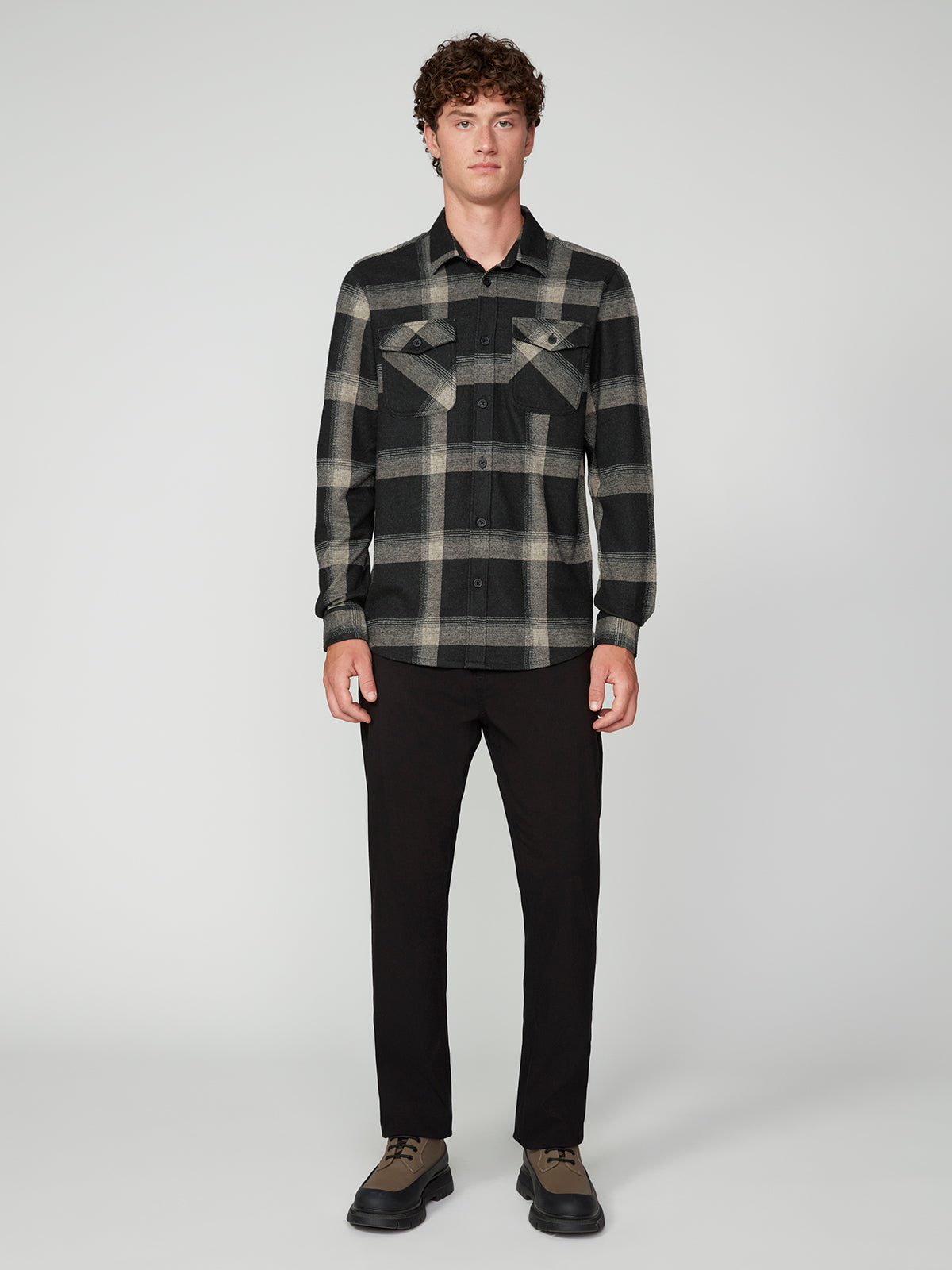 A man with curly hair wears the PROJEK RAW MENS LONG SLEEVE FLANNEL KNIT SHIRT in black and grey plaid, paired with black pants and brown lace-up shoes, standing against a plain light gray background.