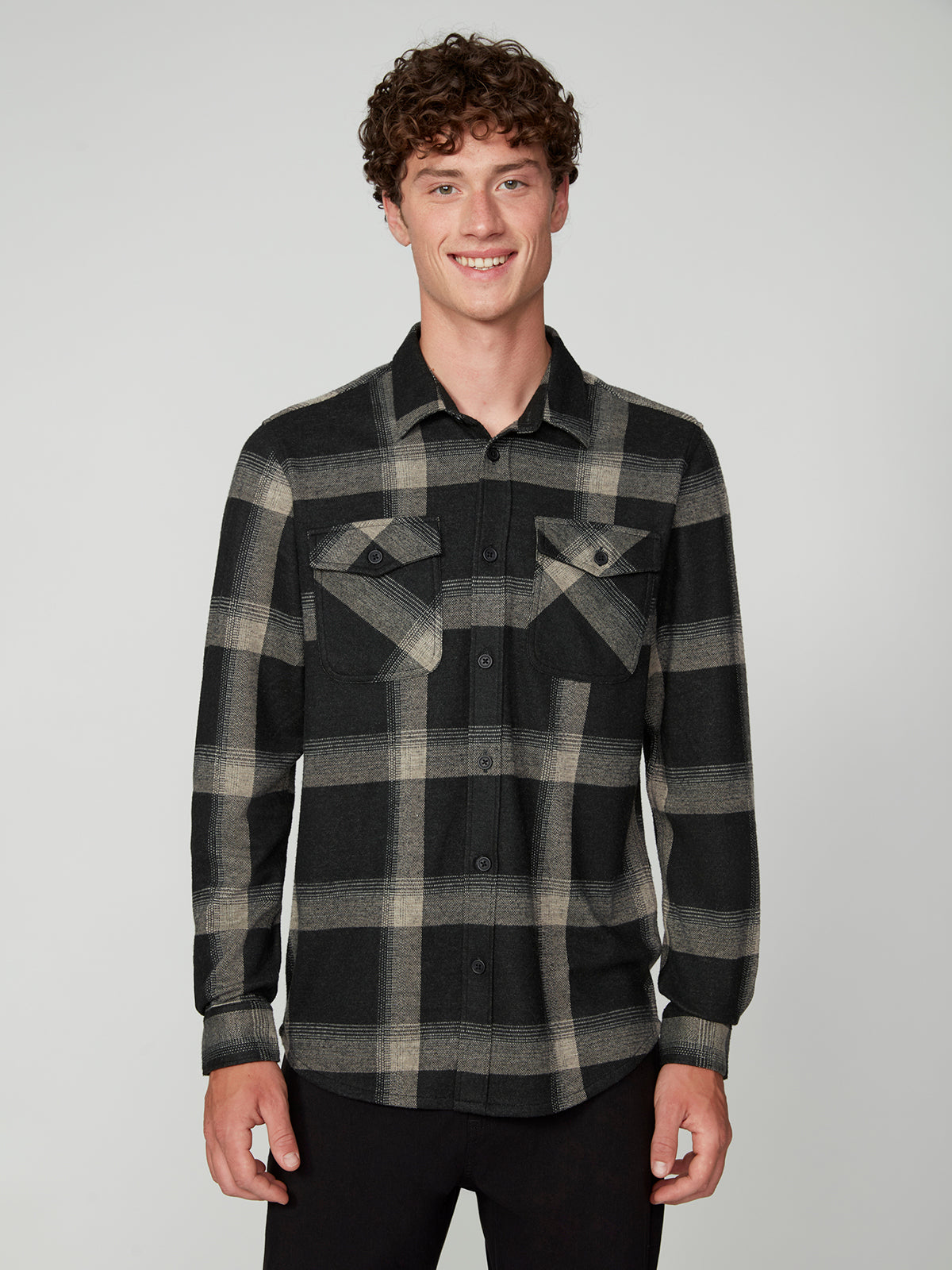 A young man with curly brown hair smiles against a plain background, wearing the PROJEK RAW MENS LONG SLEEVE FLANNEL KNIT SHIRT in black and beige plaid, featuring chest pockets, paired with black pants.
