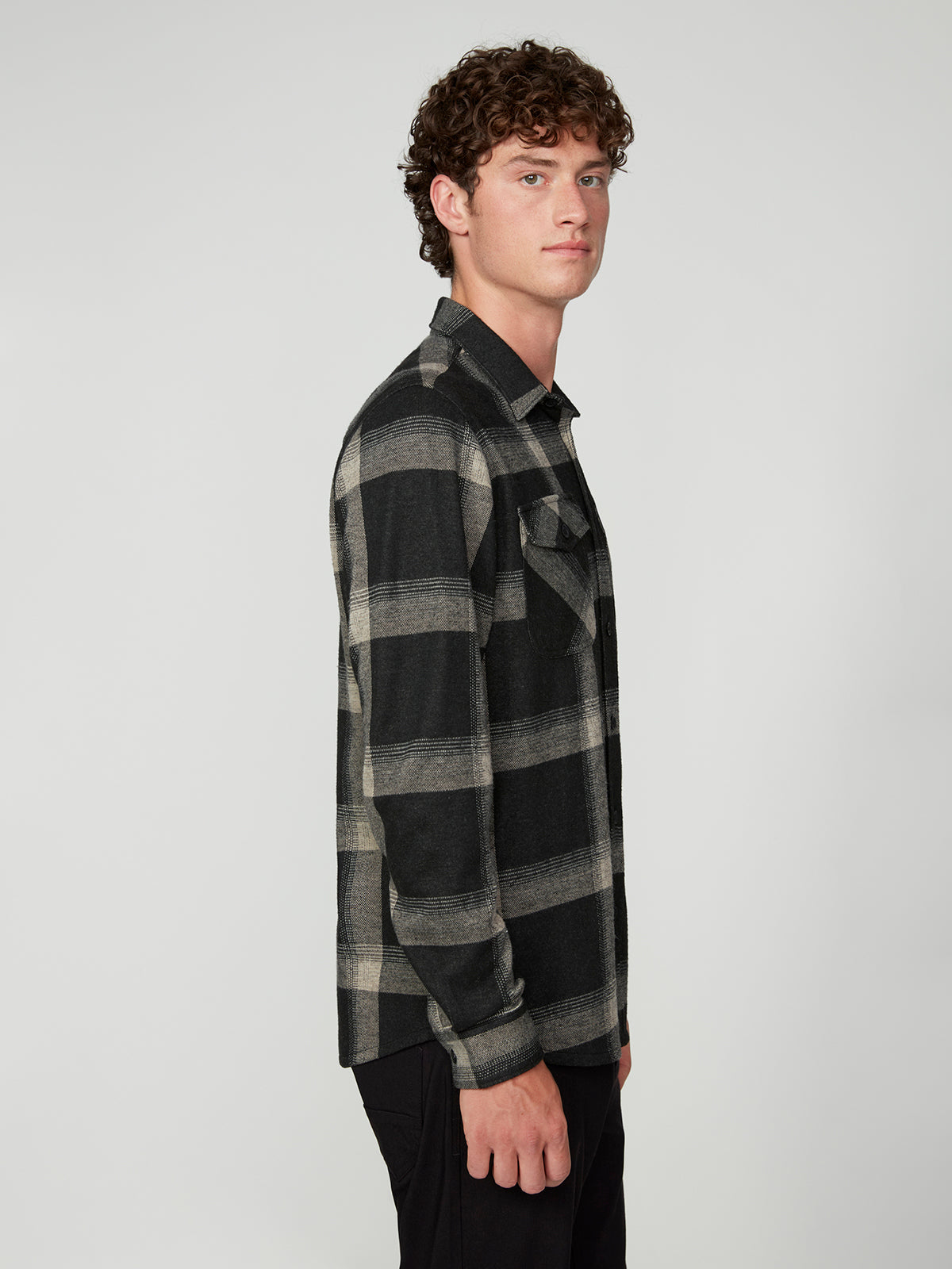 A young man with curly brown hair stands in profile, wearing a black and gray plaid PROJEK RAW MENS LONG SLEEVE FLANNEL KNIT SHIRT and black pants against a plain light gray background.