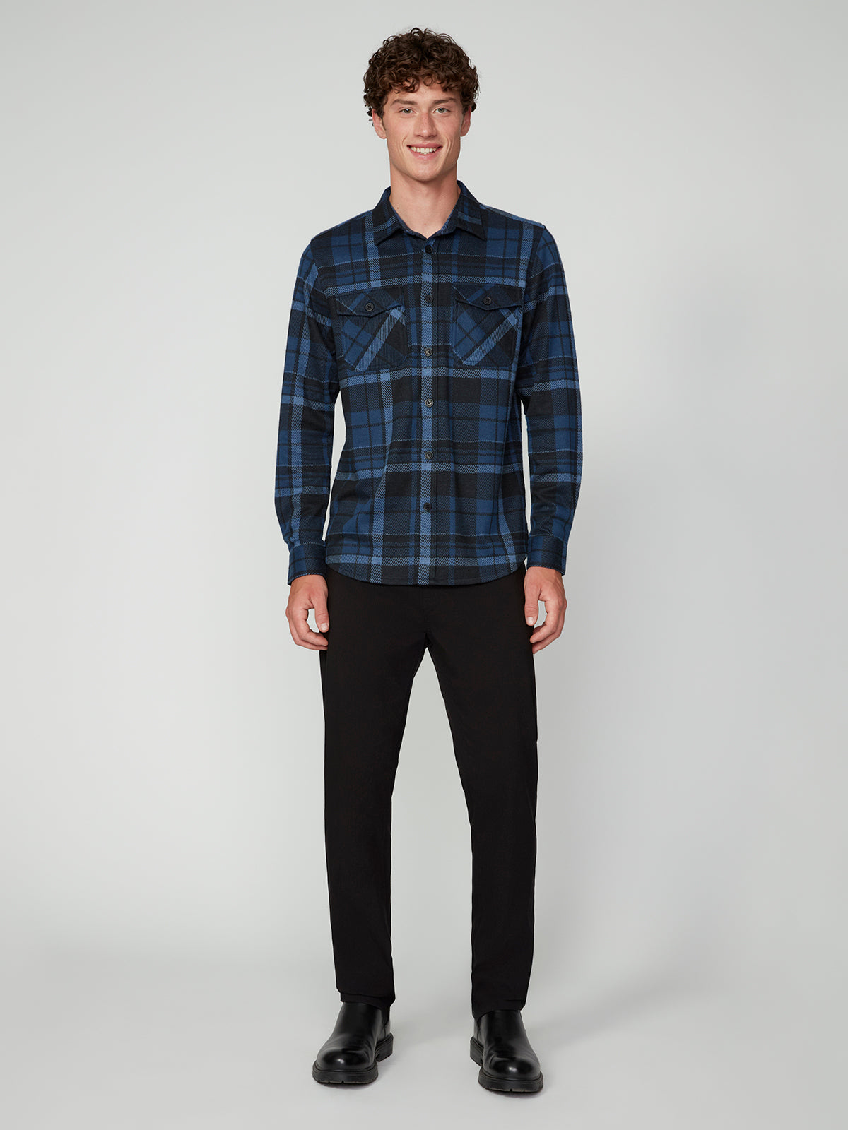 A young man with curly hair smiles, wearing a PROJEK RAW MENS LONG SLEEVE FLANNEL KNIT SHIRT in blue and black plaid, paired with black pants and boots against a plain light gray background.