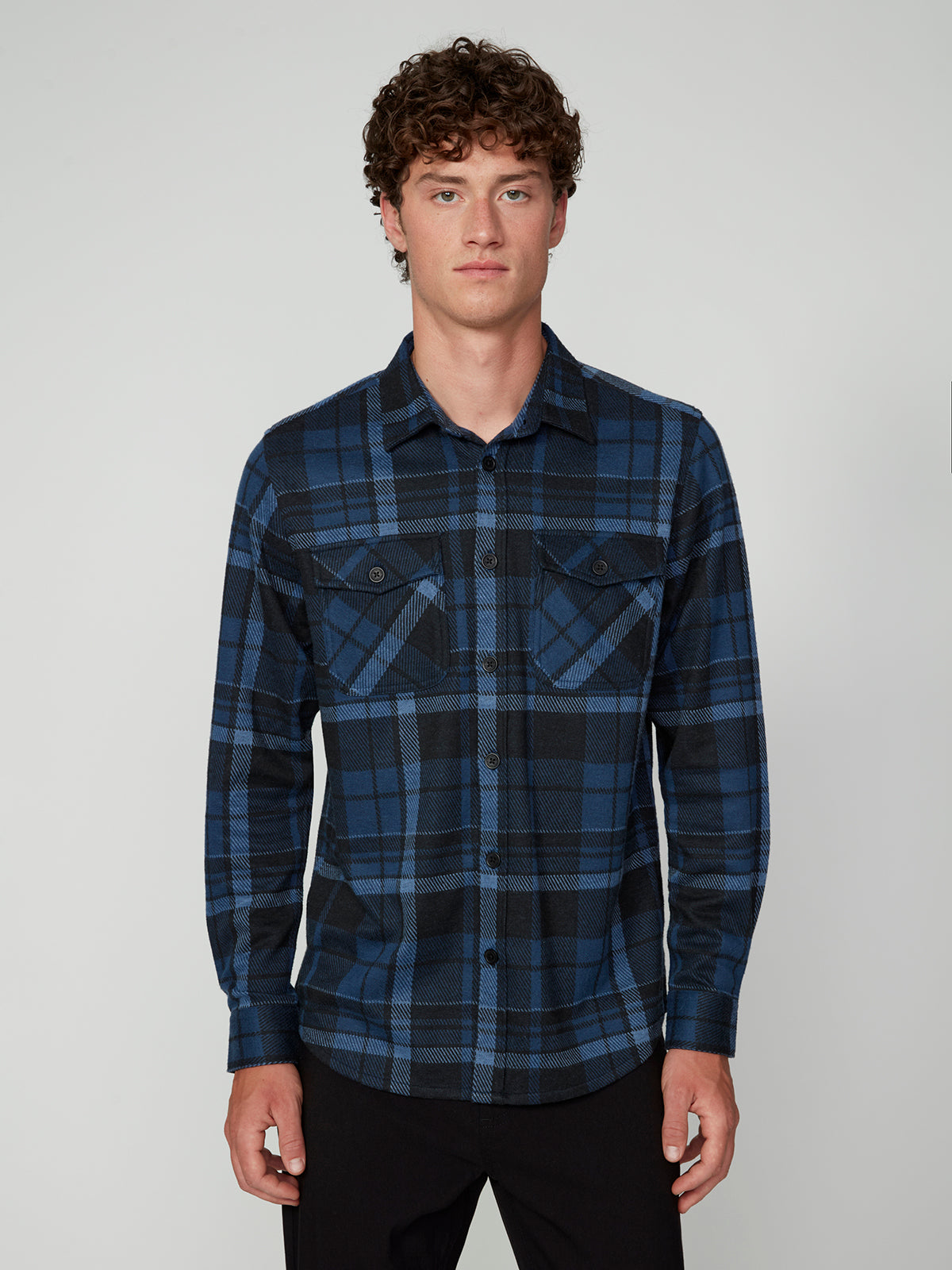 A young man with curly brown hair wears the PROJEK RAW MENS LONG SLEEVE FLANNEL KNIT SHIRT in blue and black plaid with front pockets, standing against a plain light background and looking directly at the camera with a neutral expression.