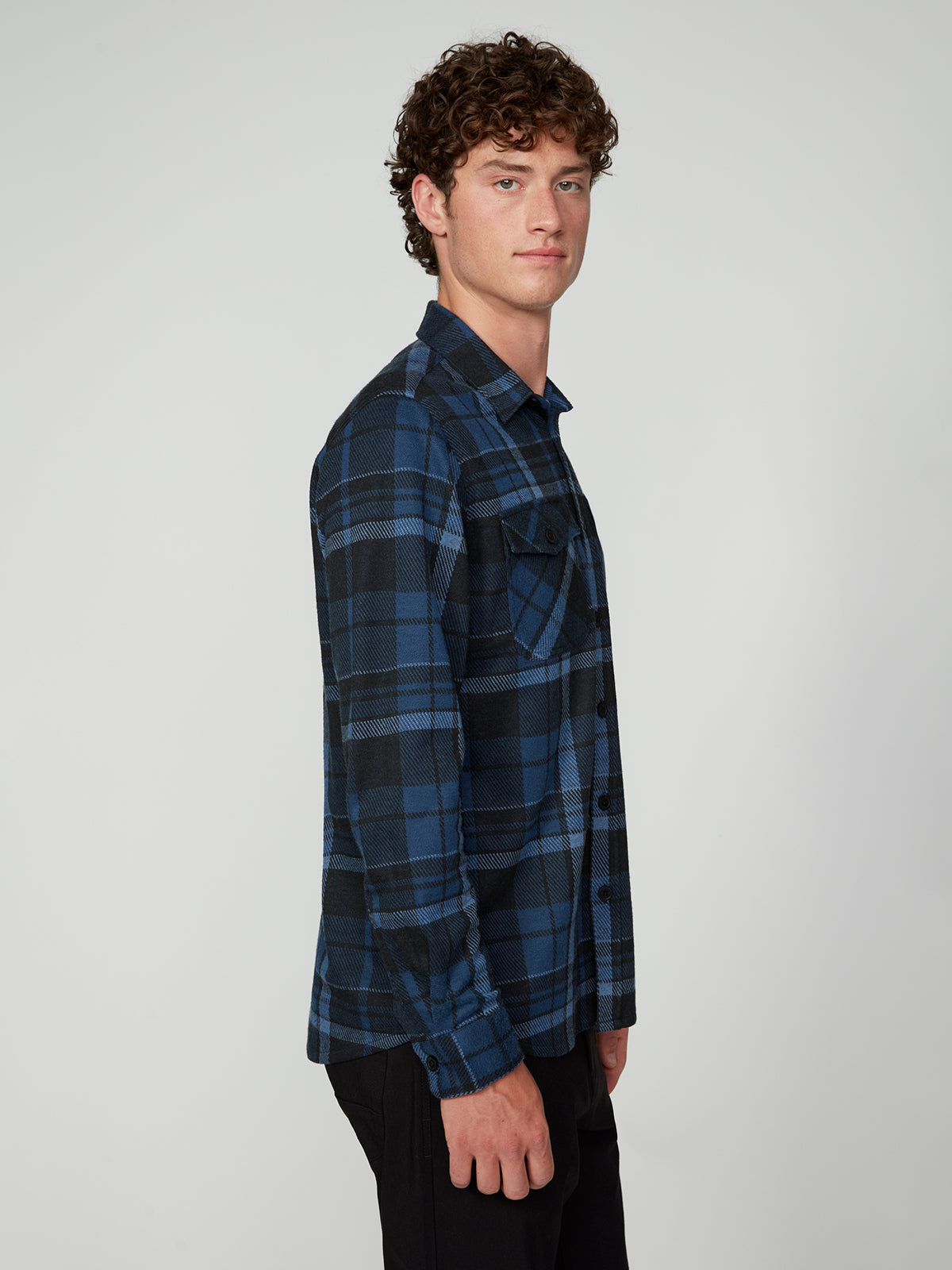 A young man with curly hair wears a PROJEK RAW MENS LONG SLEEVE FLANNEL KNIT SHIRT in blue and black plaid, standing sideways and facing the camera against a plain light gray background.