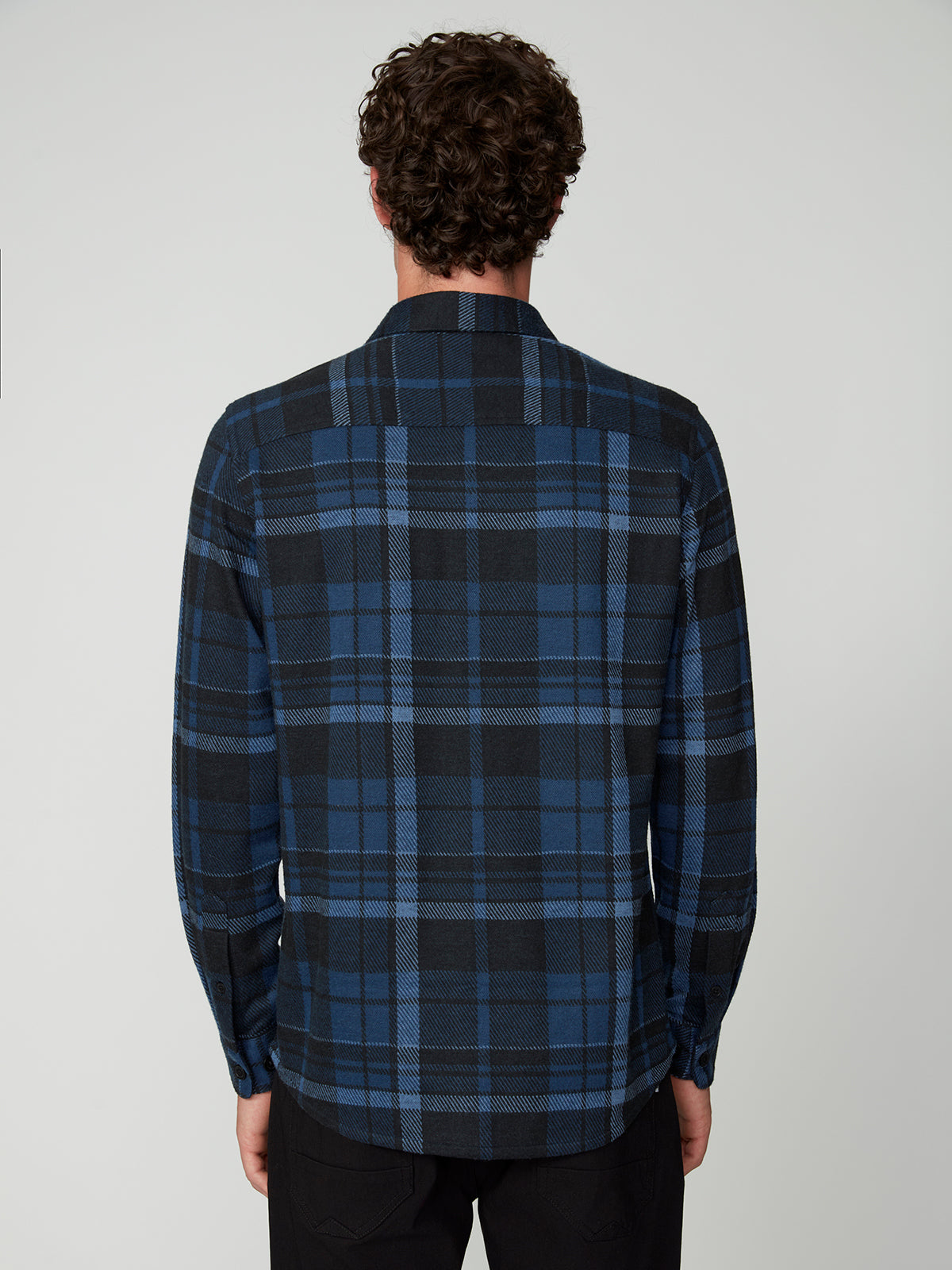 A person with curly hair is seen from behind wearing a PROJEK RAW MENS LONG SLEEVE FLANNEL KNIT SHIRT in blue and black plaid, paired with black pants, set against a simple light background.