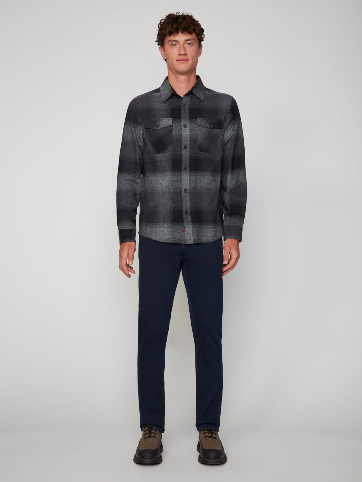 A person faces forward against a plain light gray background, wearing a PROJEK RAW MEN’S LONG SLEEVE FLANNEL SHIRT in black and gray, paired with dark blue pants and brown shoes.