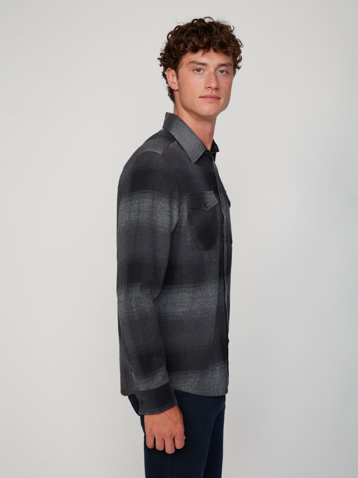 A man with curly hair stands sideways, facing left, wearing a PROJEK RAW MEN’S LONG SLEEVE FLANNEL SHIRT and dark pants against a plain light gray background.