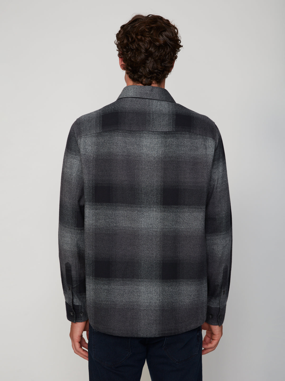 A person with short curly hair faces away from the camera, wearing the PROJEK RAW MEN’S LONG SLEEVE FLANNEL SHIRT in gray and black plaid, paired with dark pants against a plain light background.
