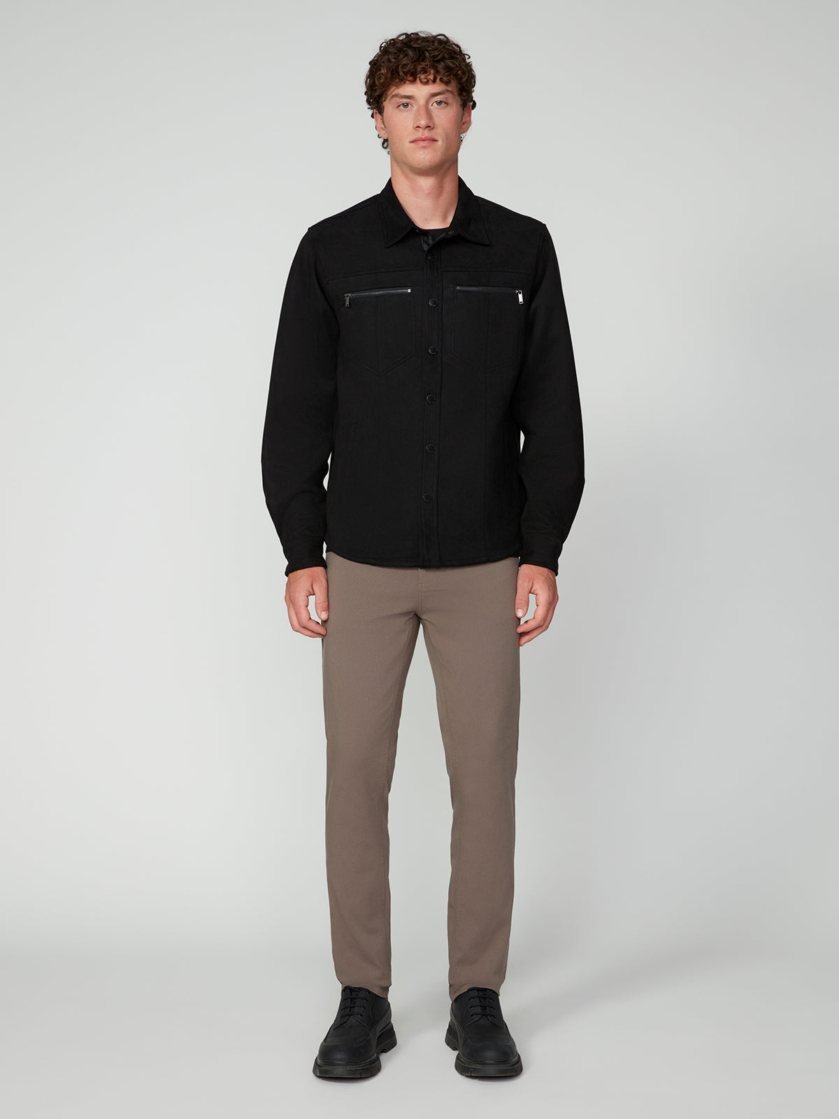A person stands facing forward against a plain background, wearing the PROJEK RAW MENS LONG SLEEVE FAUX SUEDE OVERSHIRT in black with zippered chest pockets, taupe slim-fit pants, and black shoes—a stylish lightweight layer for any occasion.