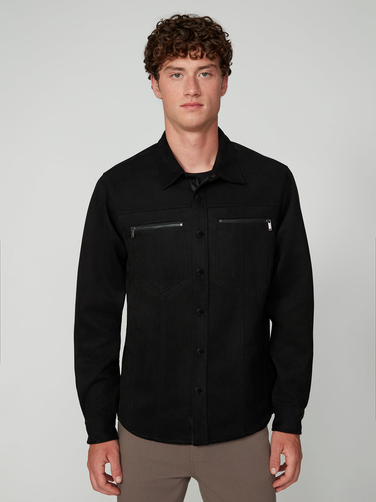 A man with short curly hair wears a black PROJEK RAW MENS LONG SLEEVE FAUX SUEDE OVERSHIRT with front zippers, standing against a plain light background.