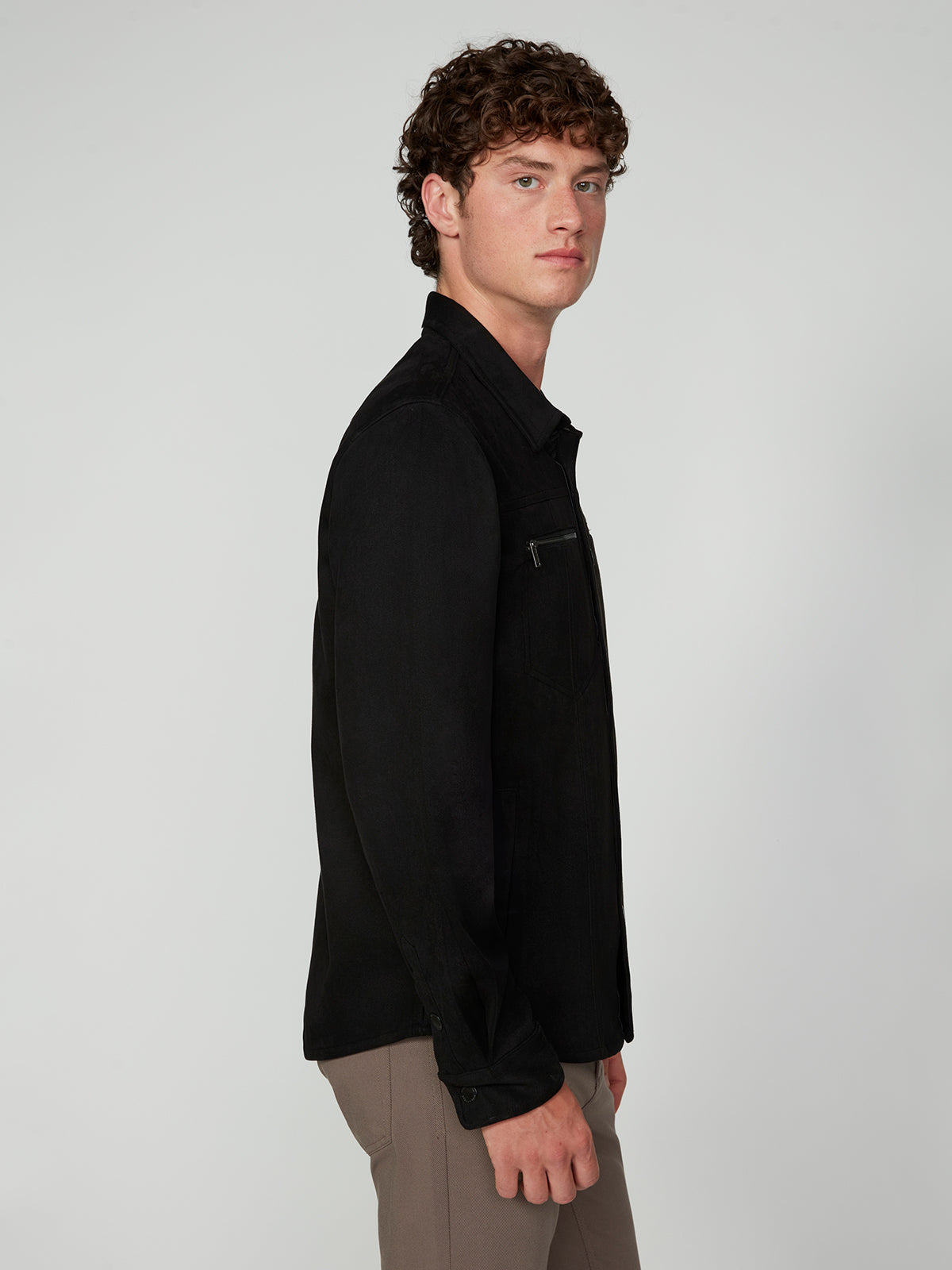 A young man with curly brown hair wears the PROJEK RAW MENS LONG SLEEVE FAUX SUEDE OVERSHIRT in black and taupe pants, posing sideways against a light gray background—the perfect lightweight layer for any outfit.