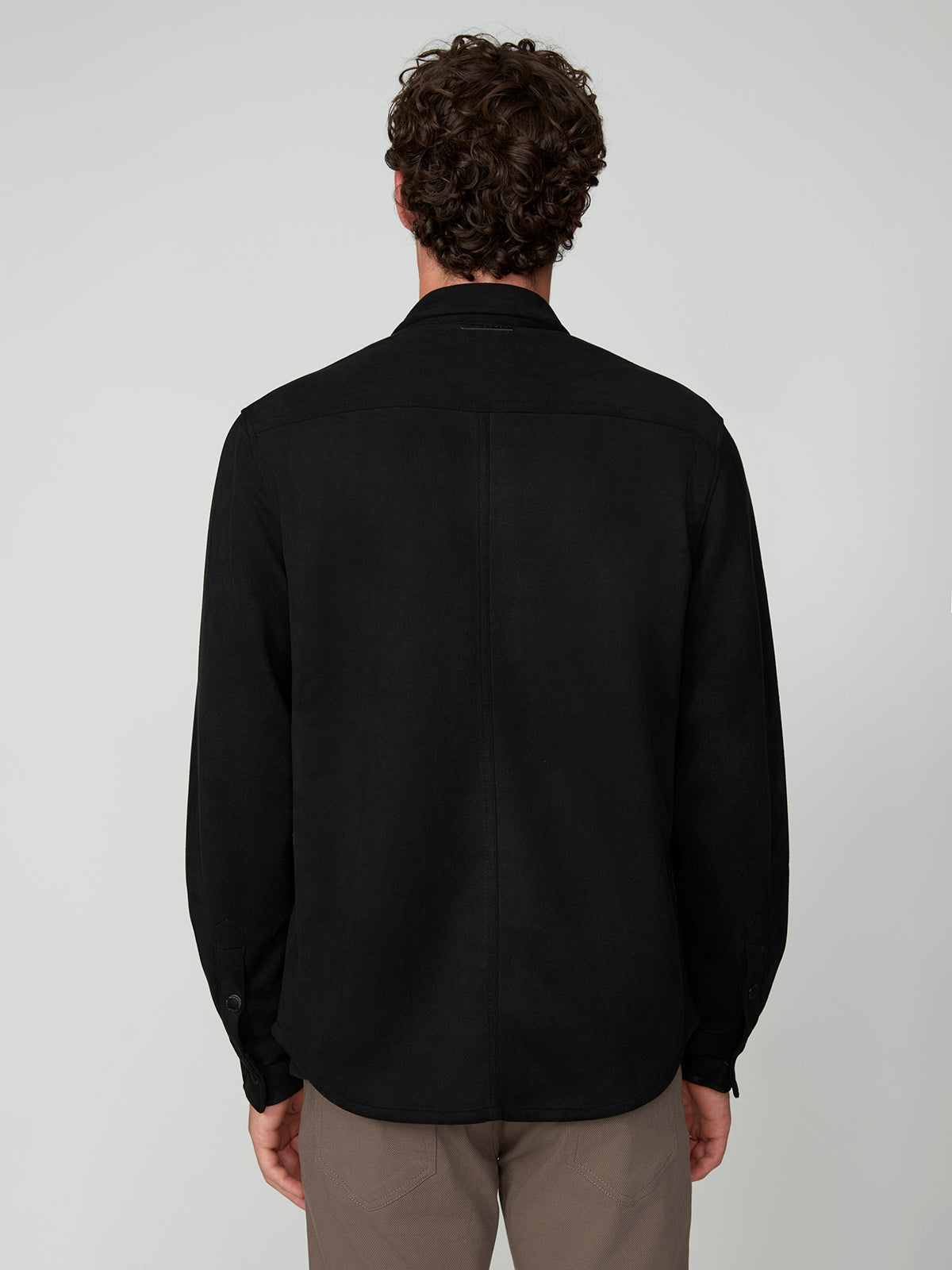 A man with curly brown hair is seen from behind, wearing the PROJEK RAW MENS LONG SLEEVE FAUX SUEDE OVERSHIRT in black and light brown pants, against a plain light gray background.