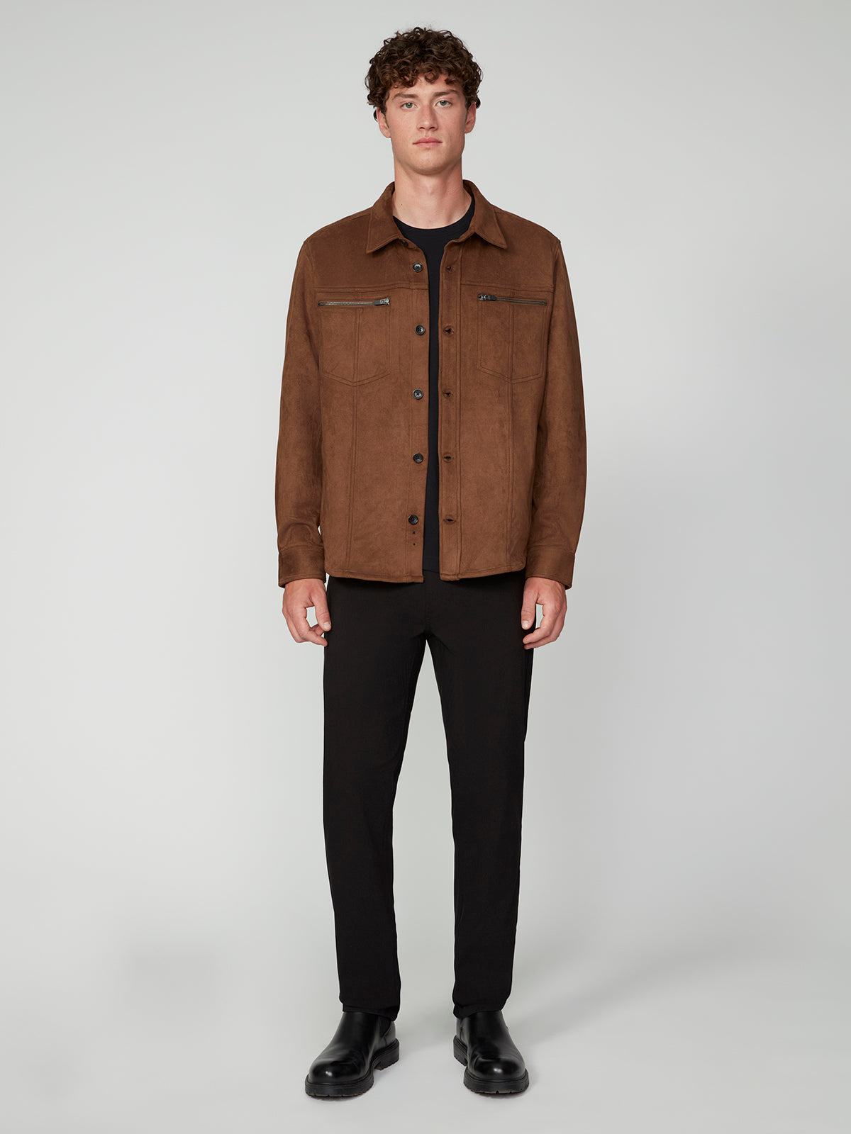 A young man with curly hair wears the PROJEK RAW MENS LONG SLEEVE FAUX SUEDE OVERSHIRT in brown over a black shirt, pants, and boots. Set against a light gray background, this piece is a perfect lightweight layer for any season.