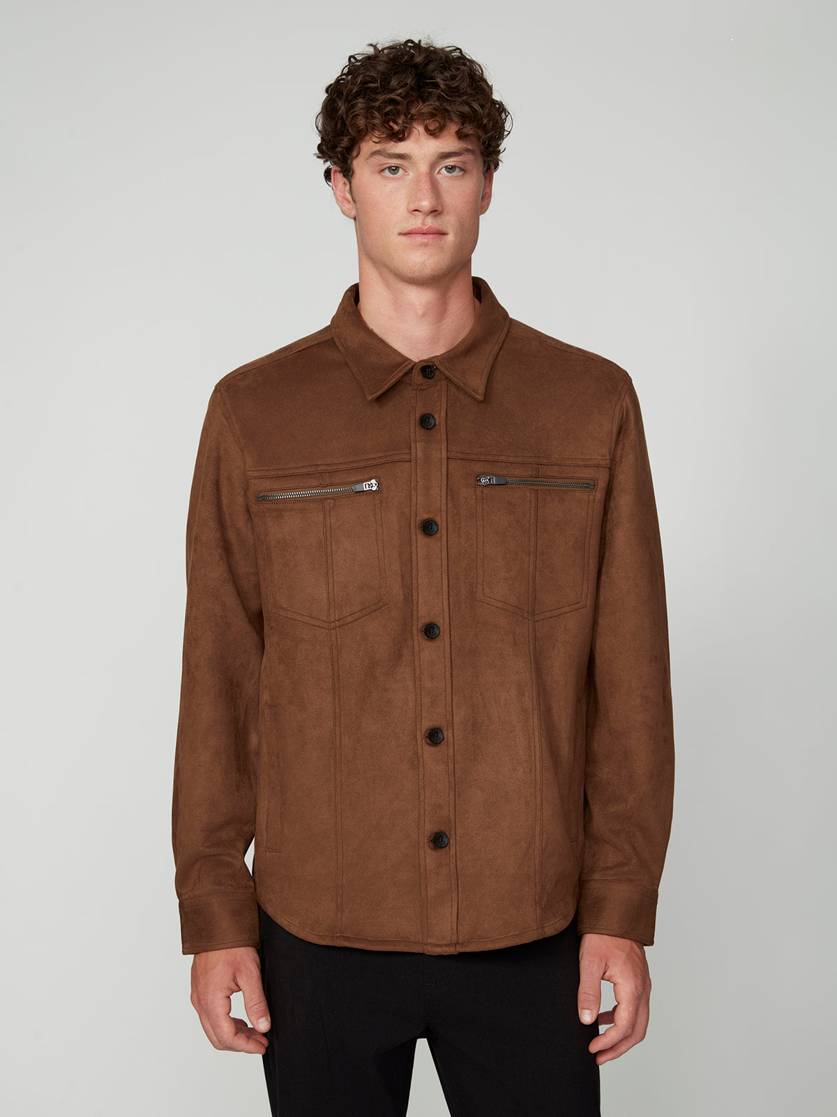 A young man with curly brown hair wears the PROJEK RAW MENS LONG SLEEVE FAUX SUEDE OVERSHIRT in brown, featuring two zippered chest pockets. He stands facing forward against a plain light gray background.
