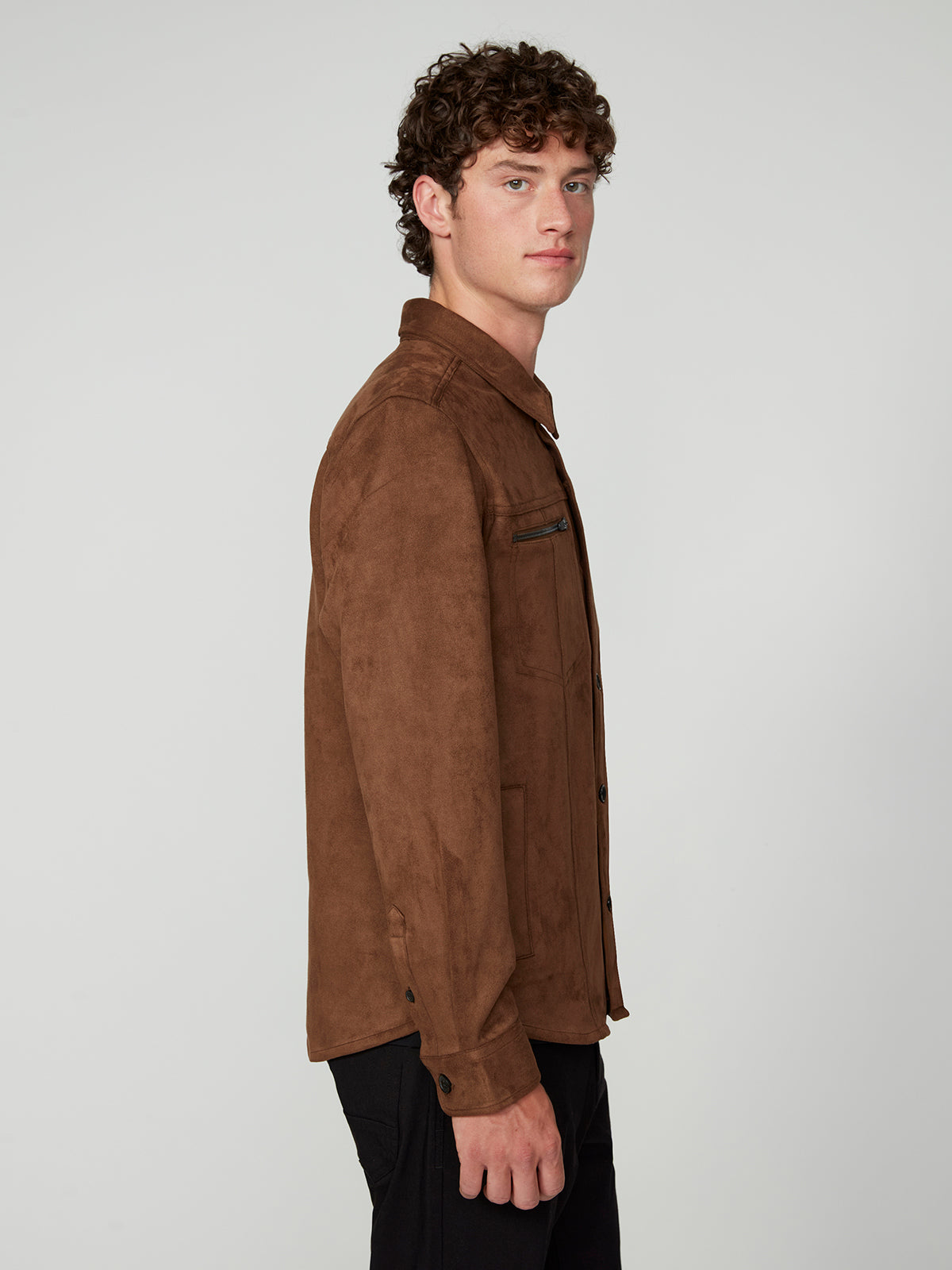 A young man with curly brown hair stands in profile, wearing the PROJEK RAW MENS LONG SLEEVE FAUX SUEDE OVERSHIRT and black pants against a plain light gray background.