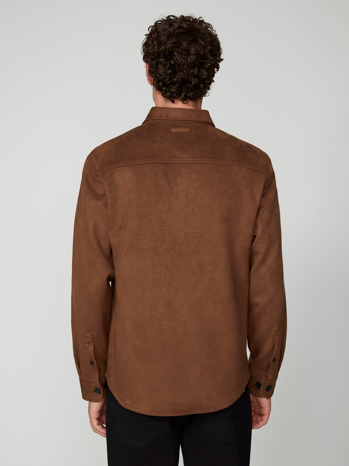 A man with curly hair, facing away from the camera, wears the PROJEK RAW MENS LONG SLEEVE FAUX SUEDE OVERSHIRT in brown and black pants against a light gray background—perfect as a lightweight layer.