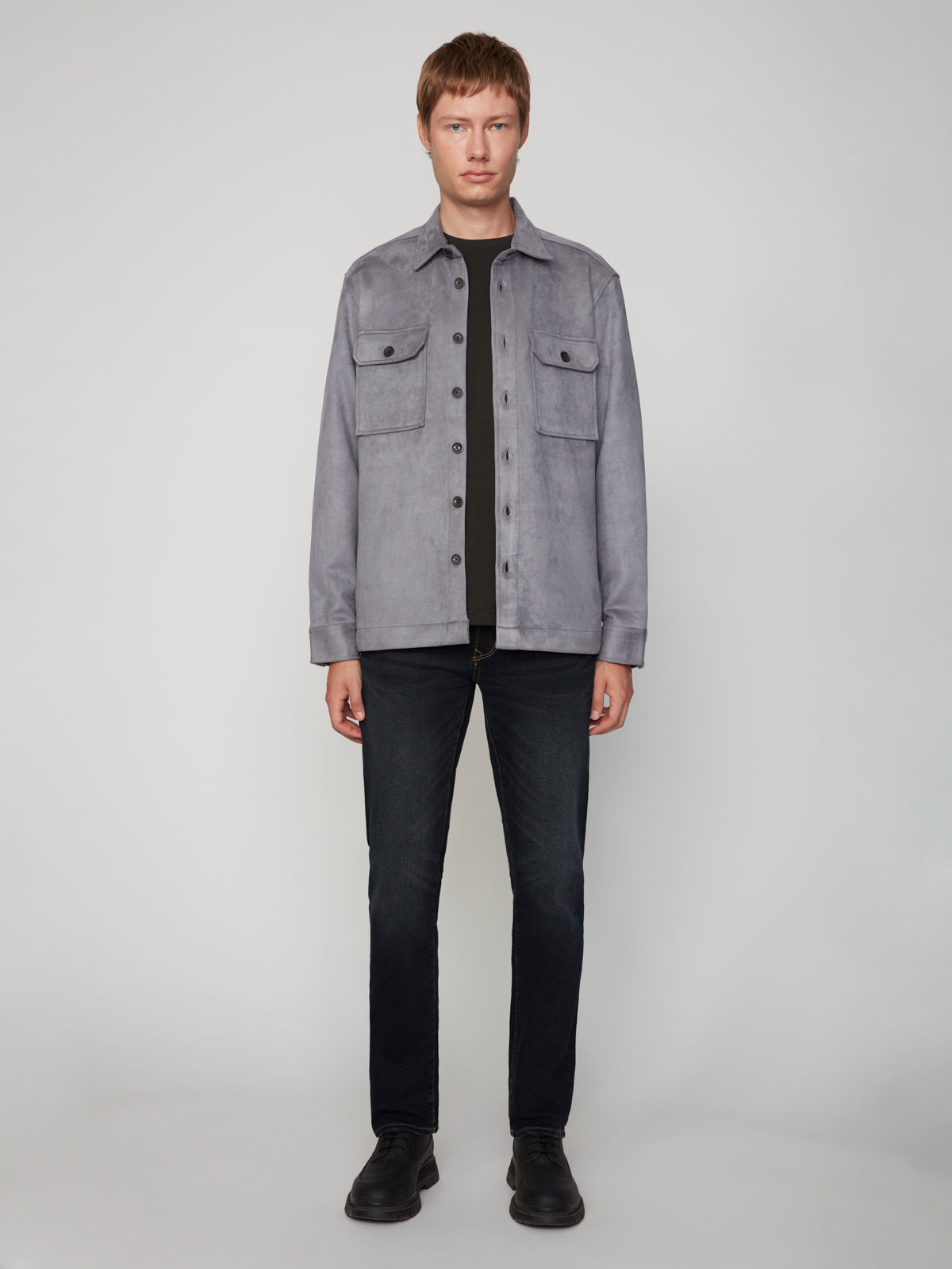 A man with short brown hair faces forward, wearing the PROJEK RAW MEN’S STRETCH FAUX SUEDE OVERSHIRT in light gray over a black shirt, paired with dark jeans and black shoes, against a plain light background.