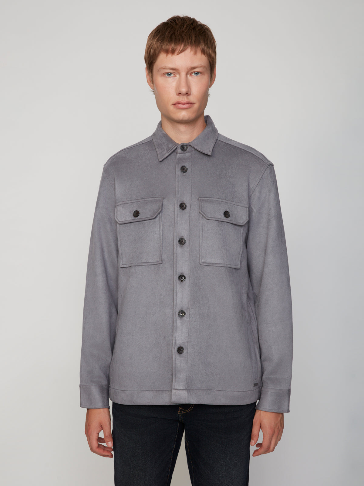 A person with short light brown hair wears the PROJEK RAW MEN’S STRETCH FAUX SUEDE OVERSHIRT in gray, featuring two chest pockets, paired with black jeans and standing against a plain light gray background.