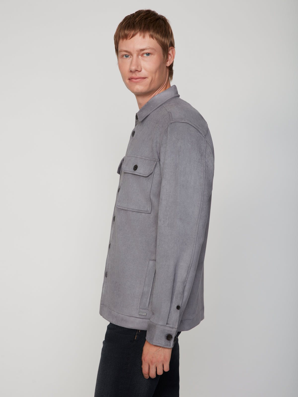 A person with short light brown hair wears the PROJEK RAW MEN’S STRETCH FAUX SUEDE OVERSHIRT in gray and dark pants, standing sideways and looking at the camera against a plain light background.