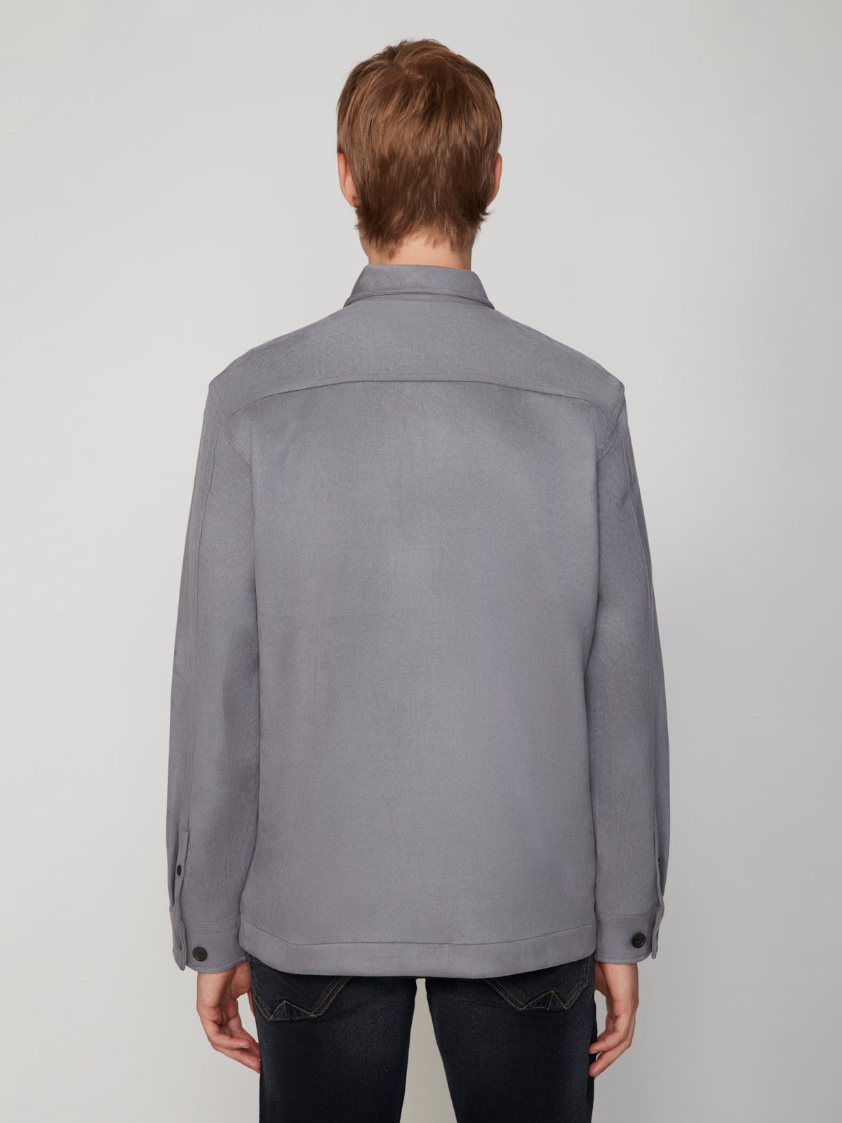 A person with short brown hair stands with their back to the camera, wearing the PROJEK RAW MEN’S STRETCH FAUX SUEDE OVERSHIRT in light gray and dark jeans against a plain light background.