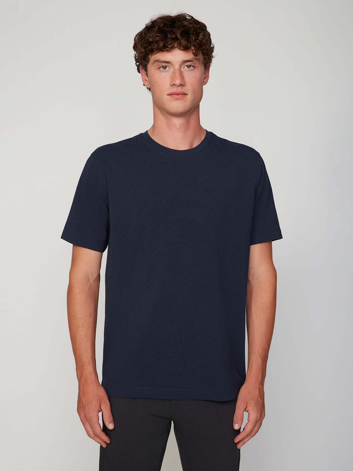 A young man with curly brown hair wears the PROJEK RAW MENS SHORT SLEEVE CREW NECK T-SHIRT in navy blue and dark pants, standing facing the camera against a light grey background—ideal for any casual wardrobe.