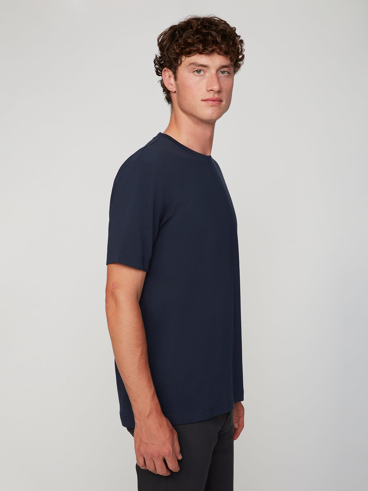 A young man with curly brown hair stands sideways, wearing a PROJEK RAW MENS SHORT SLEEVE CREW NECK T-SHIRT in navy and dark pants against a light gray background.