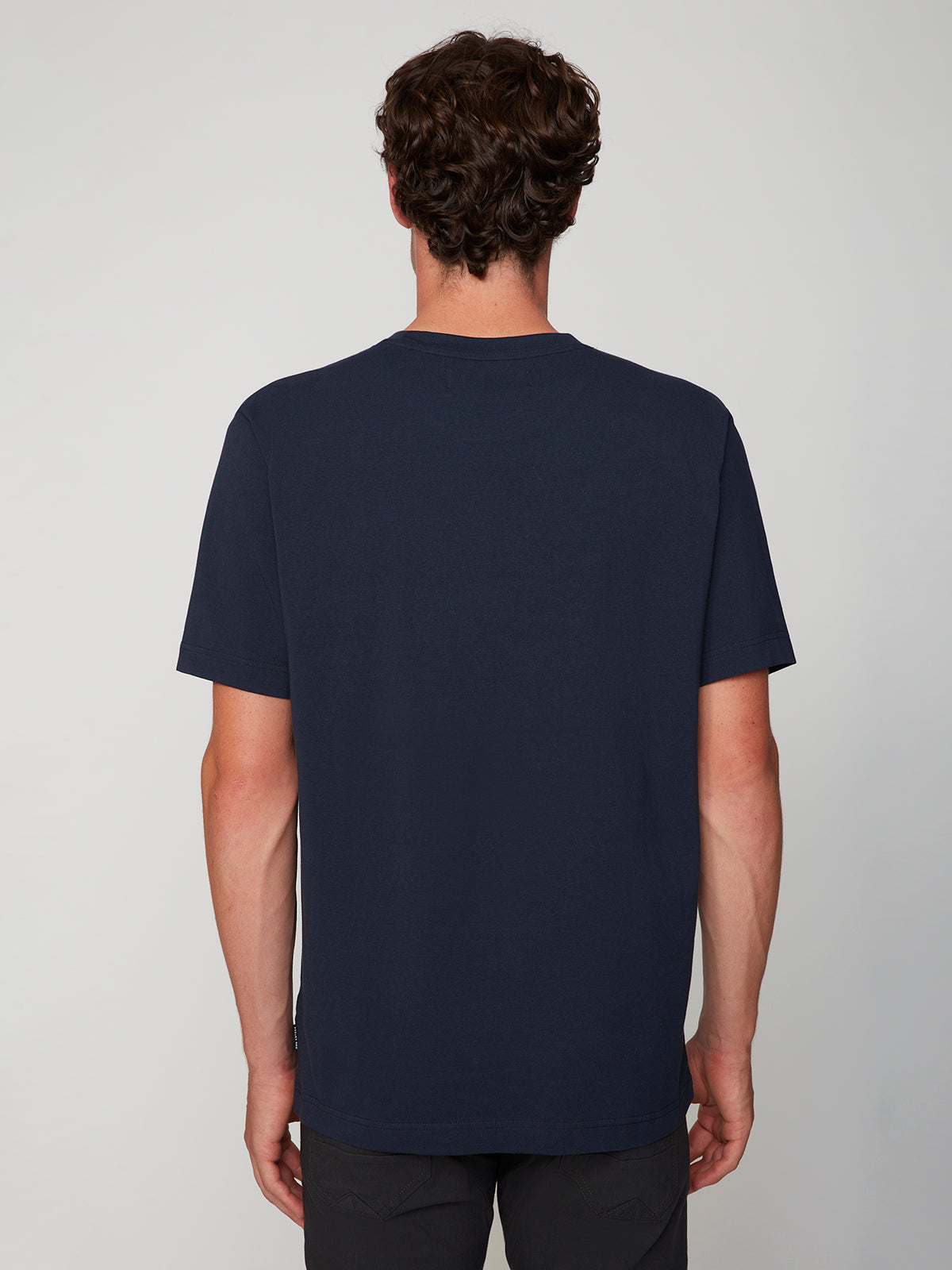 A person with short, curly brown hair faces away from the camera, modeling a PROJEK RAW MENS SHORT SLEEVE CREW NECK T-SHIRT in navy blue and dark pants against a plain light gray background—a great choice for any casual look.