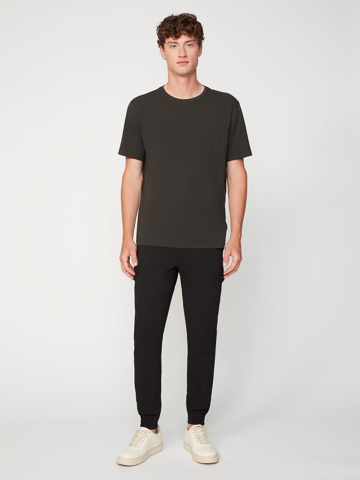 A young man faces forward against a plain white background, wearing the PROJEK RAW MENS SHORT SLEEVE CREW NECK T-SHIRT, black joggers, and white sneakers—an essential casual wardrobe look.