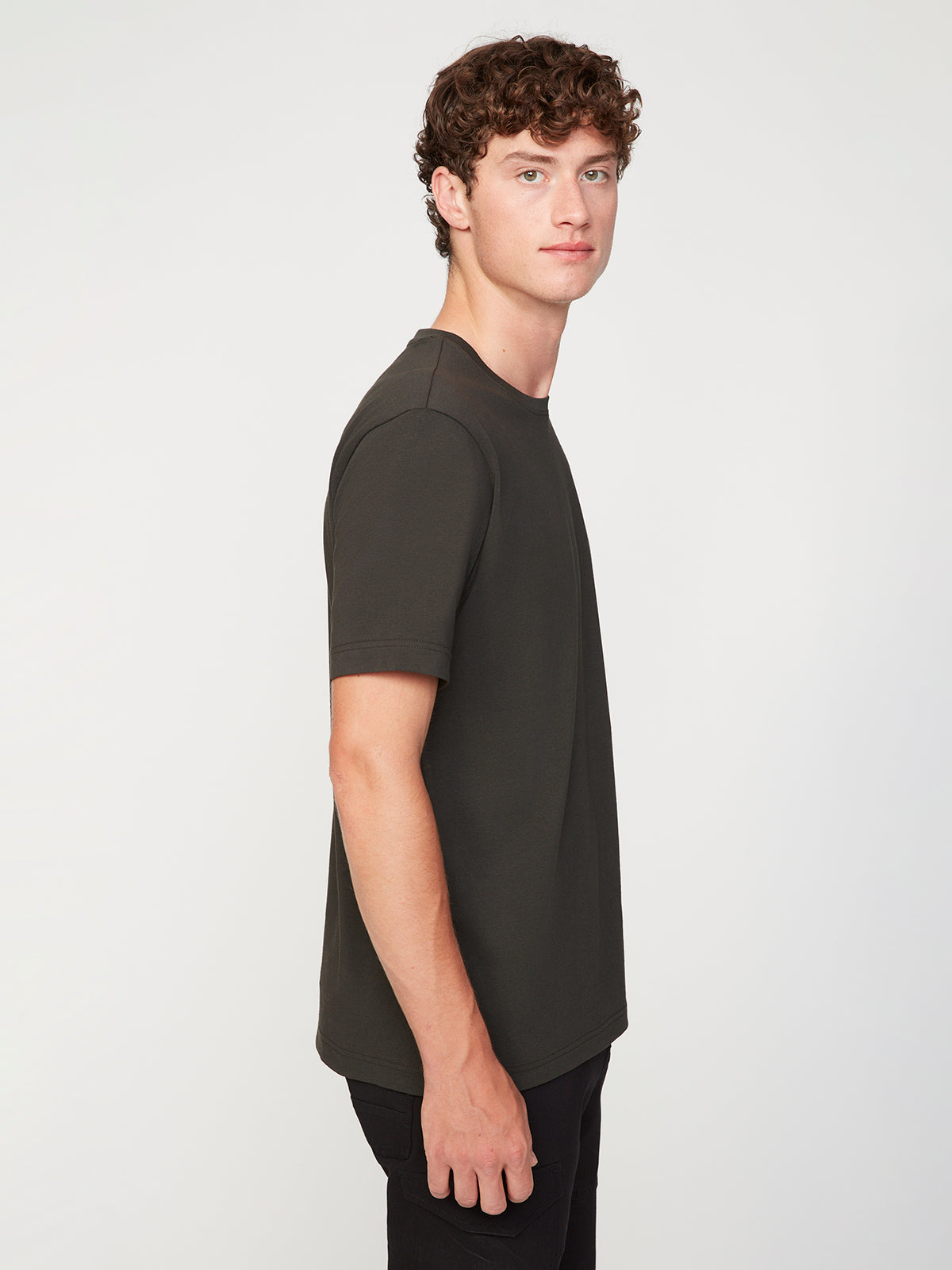 A young man with curly brown hair stands sideways, wearing a PROJEK RAW MENS SHORT SLEEVE CREW NECK T-SHIRT and black pants. He poses casually with a neutral expression against a plain white background.