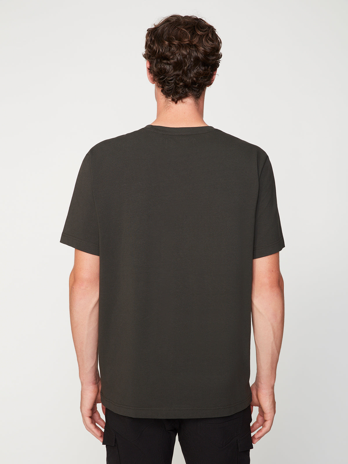 A man with short, curly brown hair is seen from the back wearing the PROJEK RAW MENS SHORT SLEEVE CREW NECK T-SHIRT and black pants against a plain white background—an essential for any casual wardrobe.