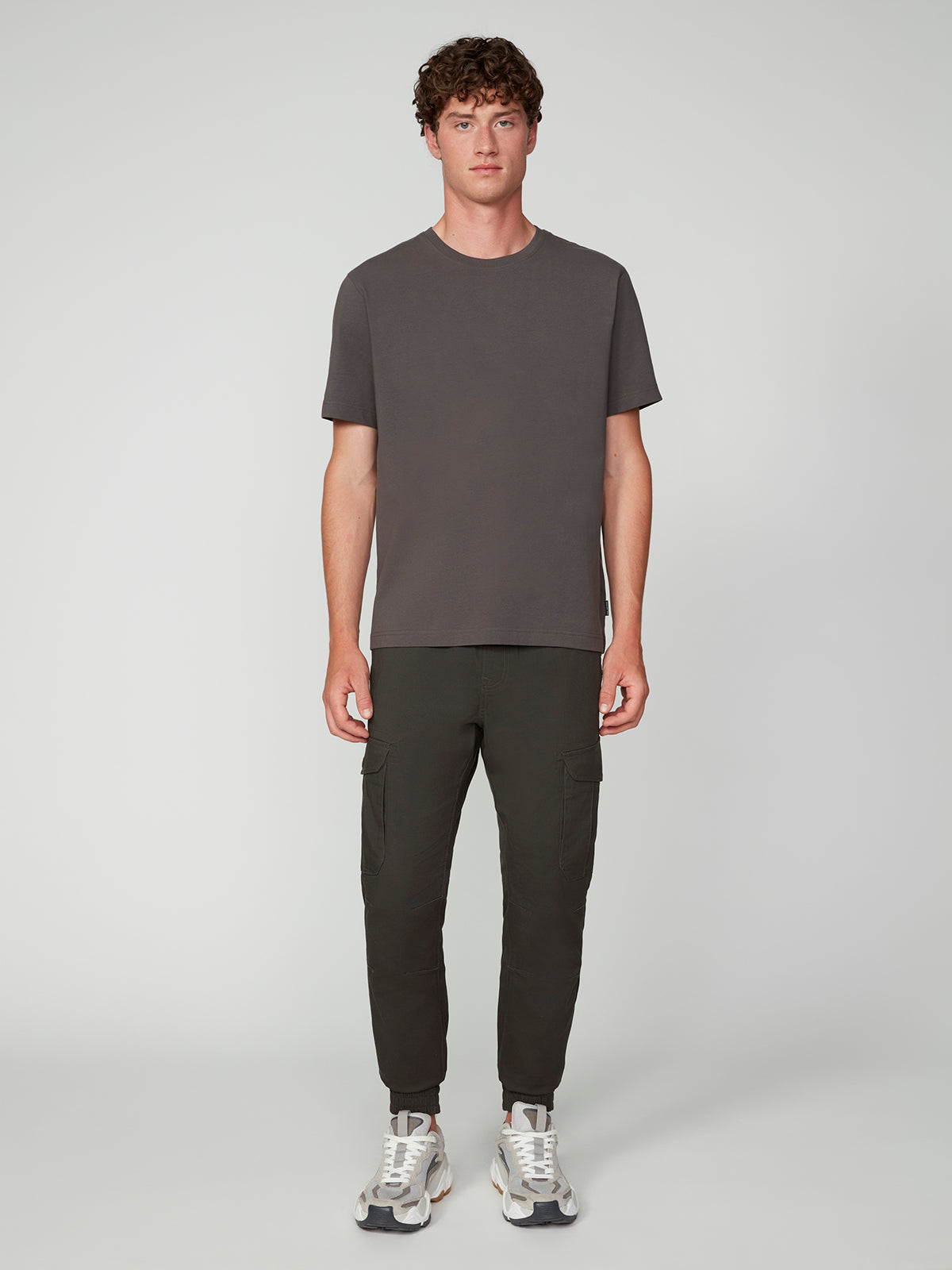 A young man with curly hair faces forward, wearing a PROJEK RAW MENS SHORT SLEEVE CREW NECK T-SHIRT in dark gray, dark jogger pants with side pockets, and white sneakers—creating a perfect casual look against a plain light background.