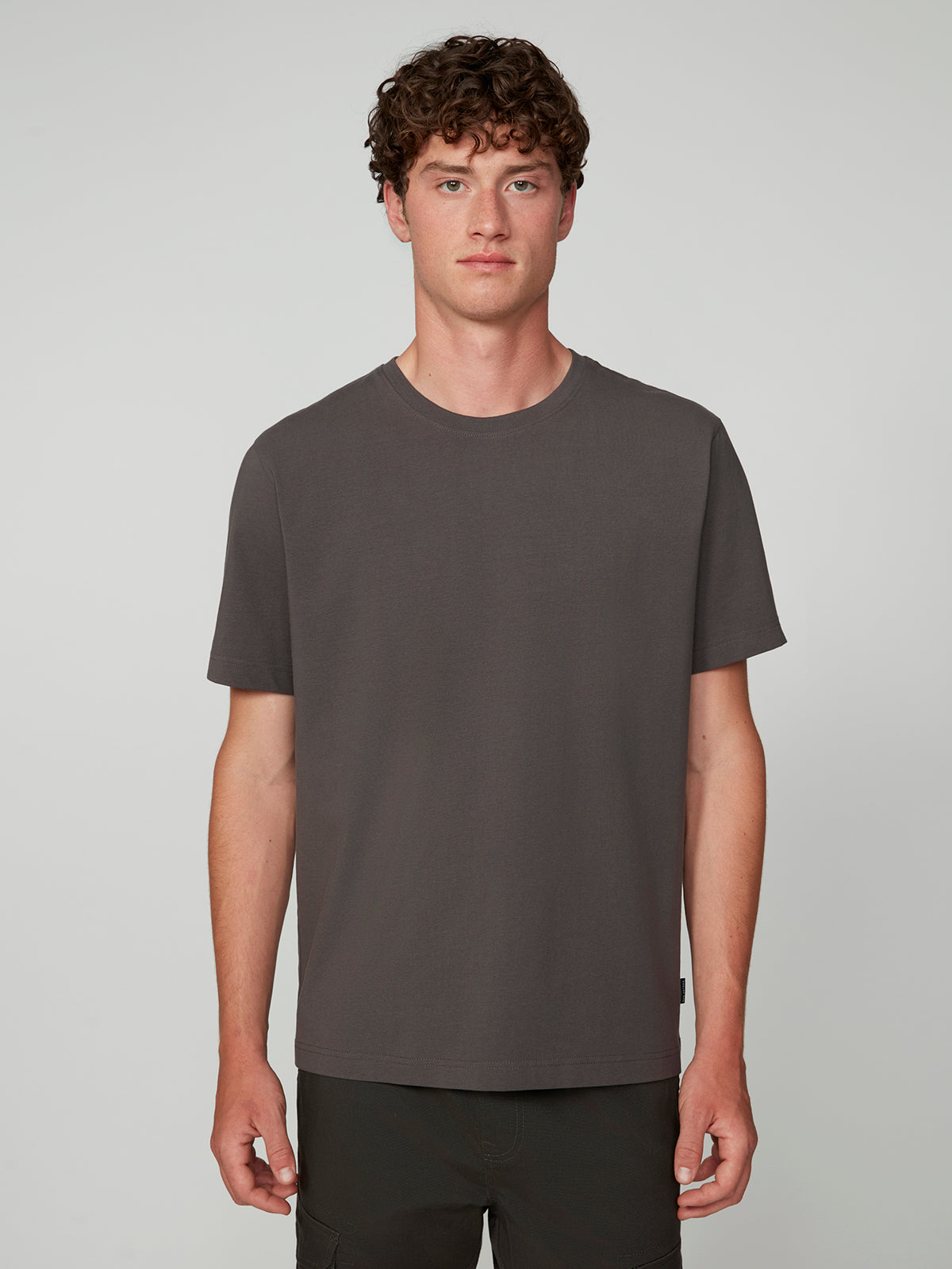 A young man with short curly hair models the PROJEK RAW MENS SHORT SLEEVE CREW NECK T-SHIRT in dark gray, paired with black pants against a light gray background—an essential piece for any casual wardrobe.