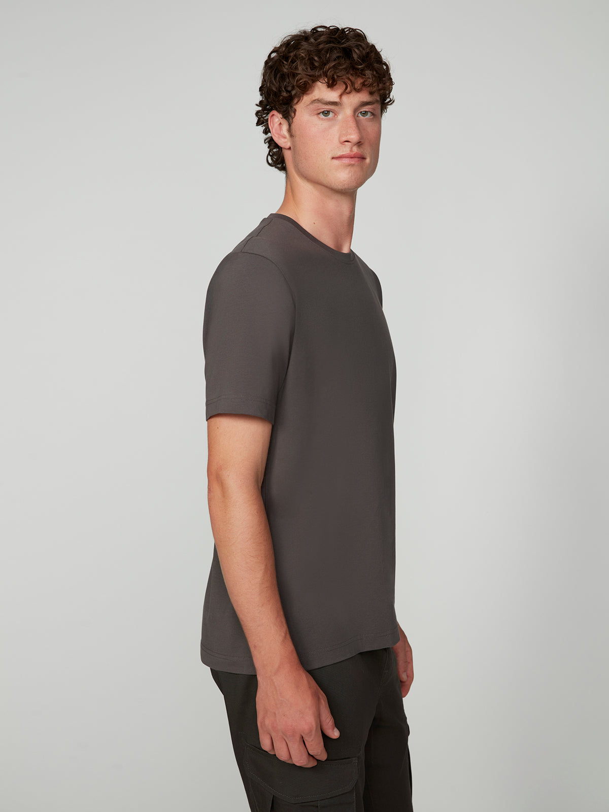 A young man with curly brown hair stands sideways, calmly facing the camera in a PROJEK RAW MENS SHORT SLEEVE CREW NECK T-SHIRT and dark pants against a light gray studio background.
