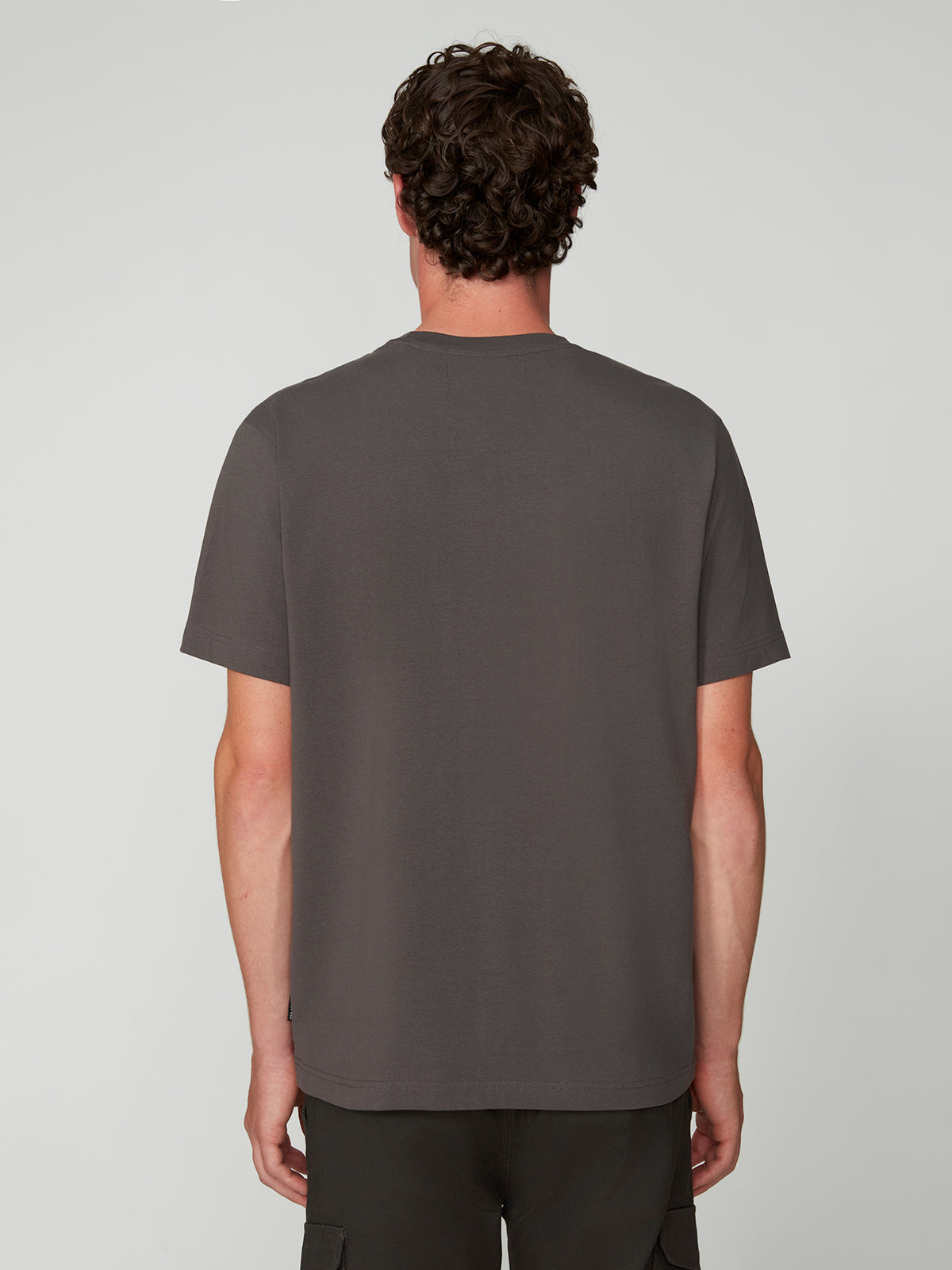 A man with curly brown hair faces away from the camera, wearing a PROJEK RAW MENS SHORT SLEEVE CREW NECK T-SHIRT in dark gray and dark pants, standing against a light gray background.