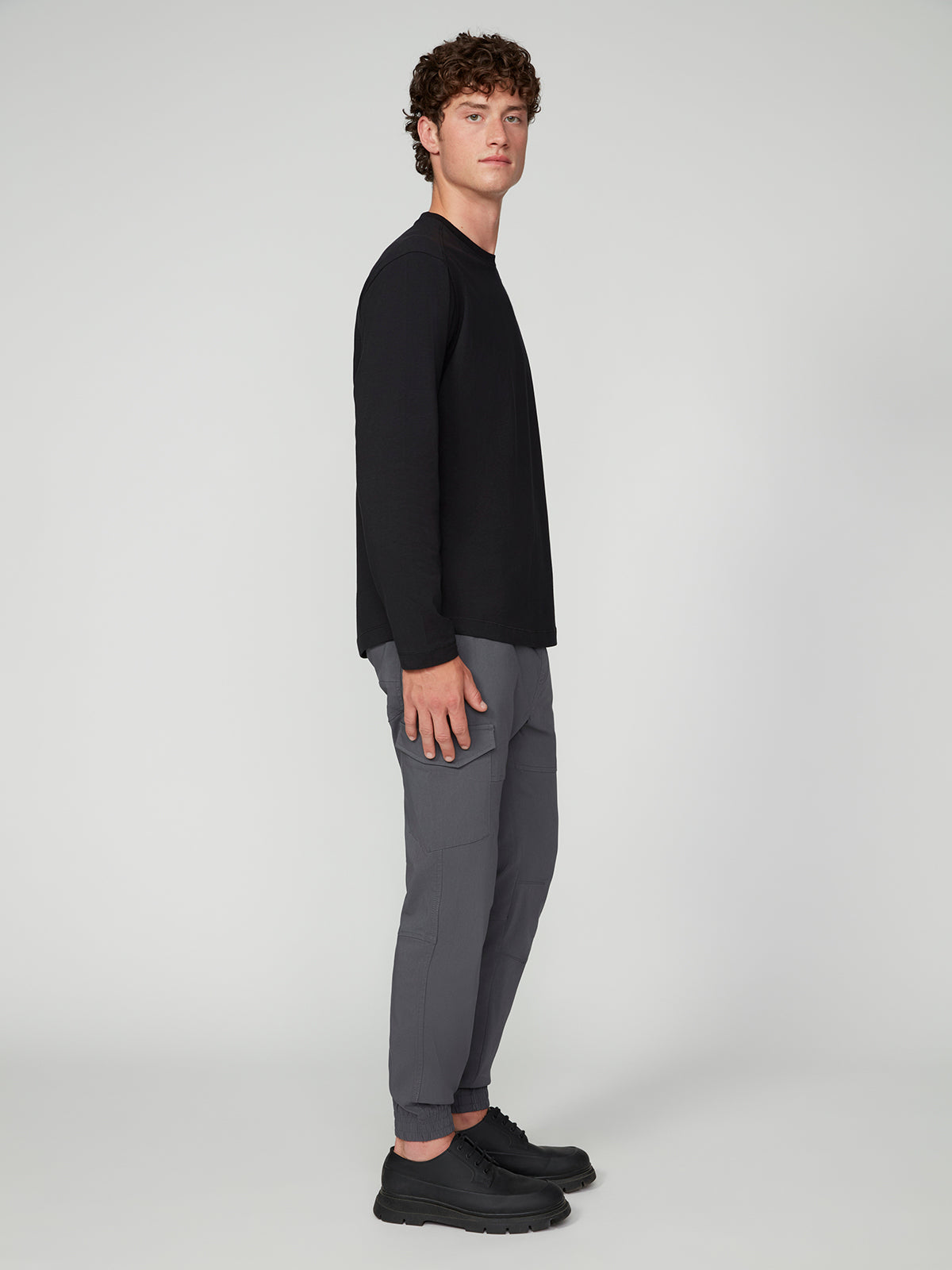 A young man with curly hair stands sideways, looking at the camera. He wears the PROJEK RAW 147333 BLACK long-sleeve shirt with gray pants and black shoes against a plain light background.