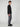 A young man with curly hair stands sideways, looking at the camera. He wears the PROJEK RAW 147333 BLACK long-sleeve shirt with gray pants and black shoes against a plain light background.