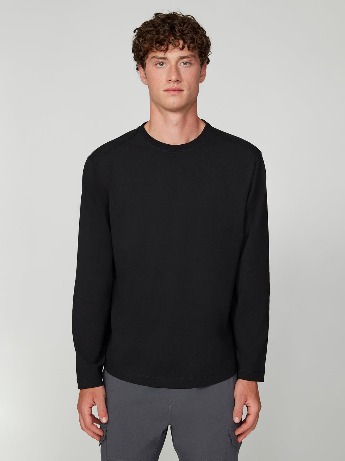 A young man with curly brown hair, wearing the PROJEK RAW 147333 BLACK long-sleeve shirt and gray pants, stands facing forward against a light gray background.