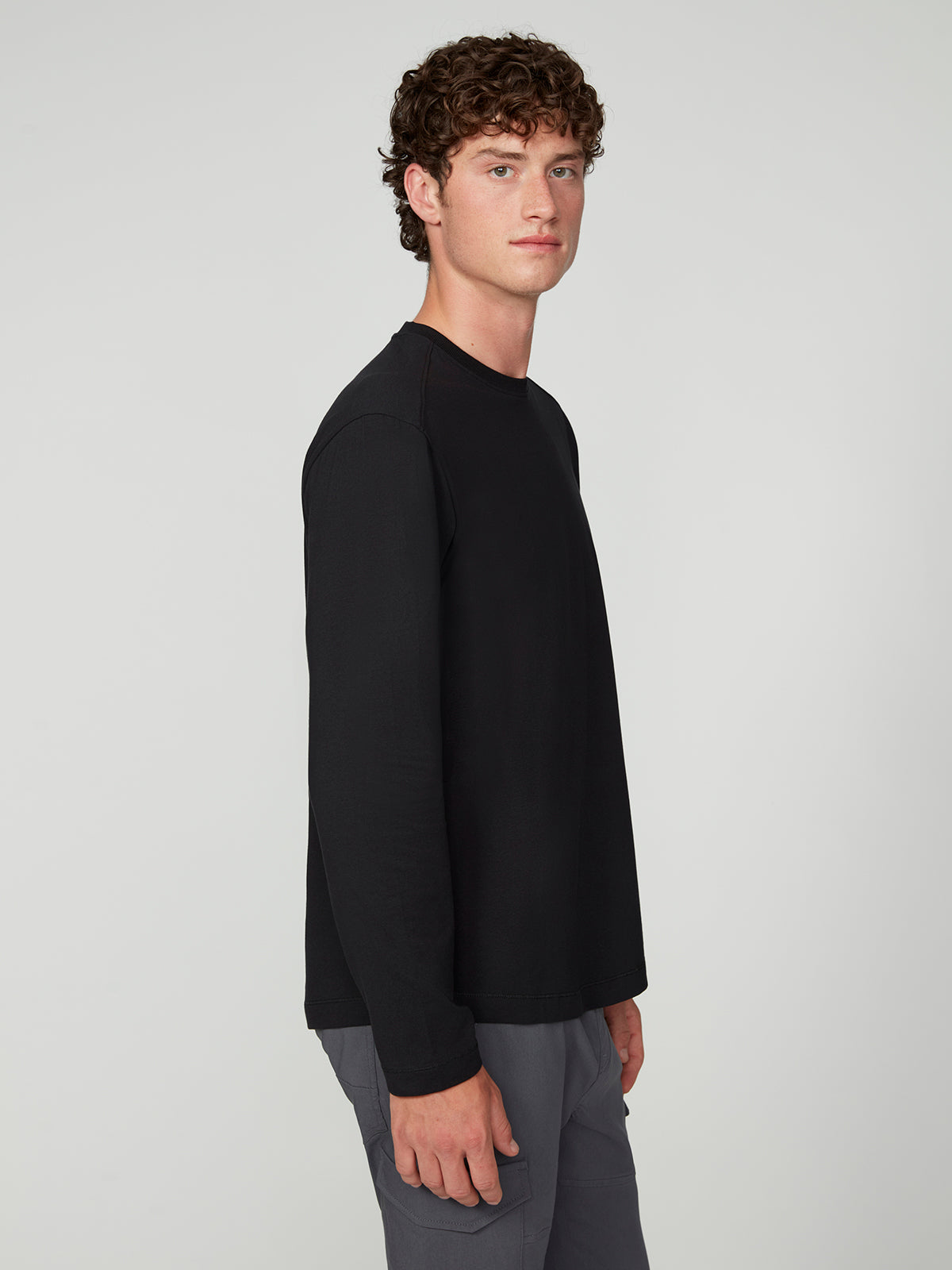 A young man with curly brown hair stands sideways, looking at the camera against a plain white background, wearing a PROJEK RAW 147333 BLACK long-sleeve shirt and gray pants.