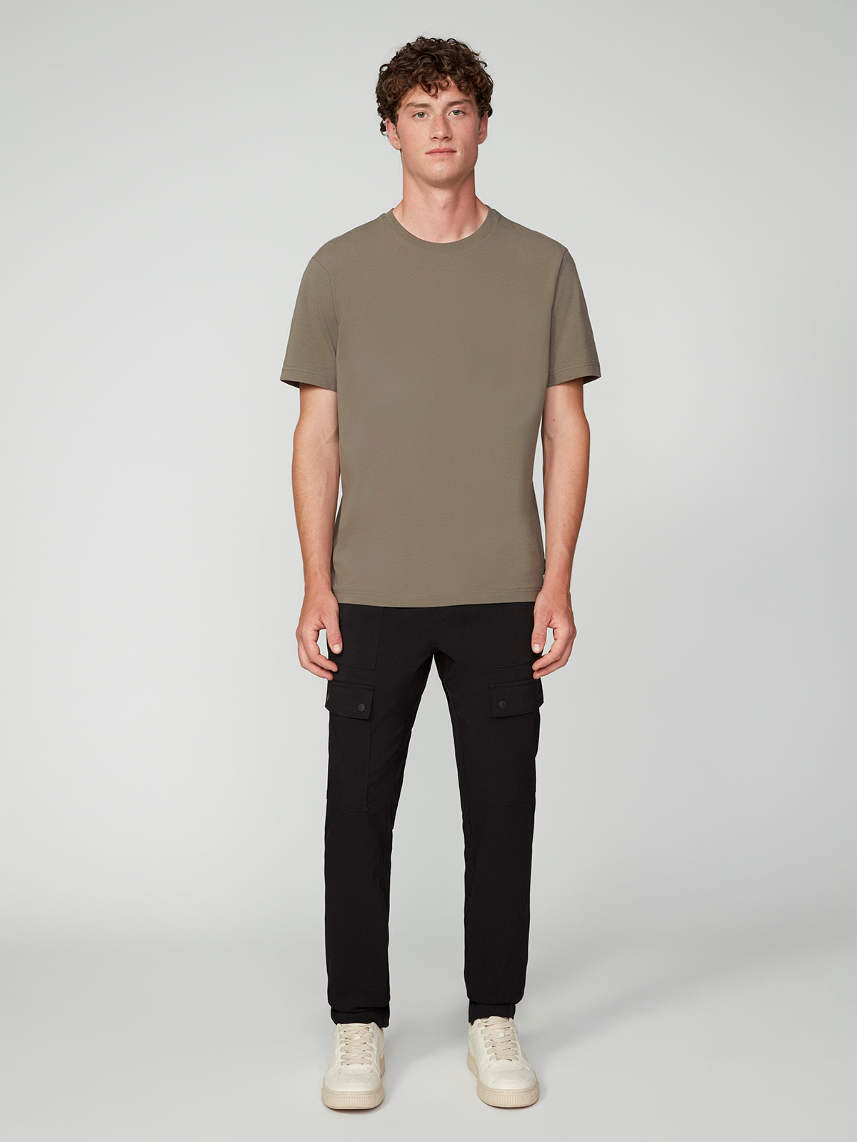 A young man with short curly hair stands facing forward in a simple gray setting, wearing a PROJEK RAW MENS SHORT SLEEVE CREW NECK T-SHIRT in olive green, black cargo pants, and white sneakers.