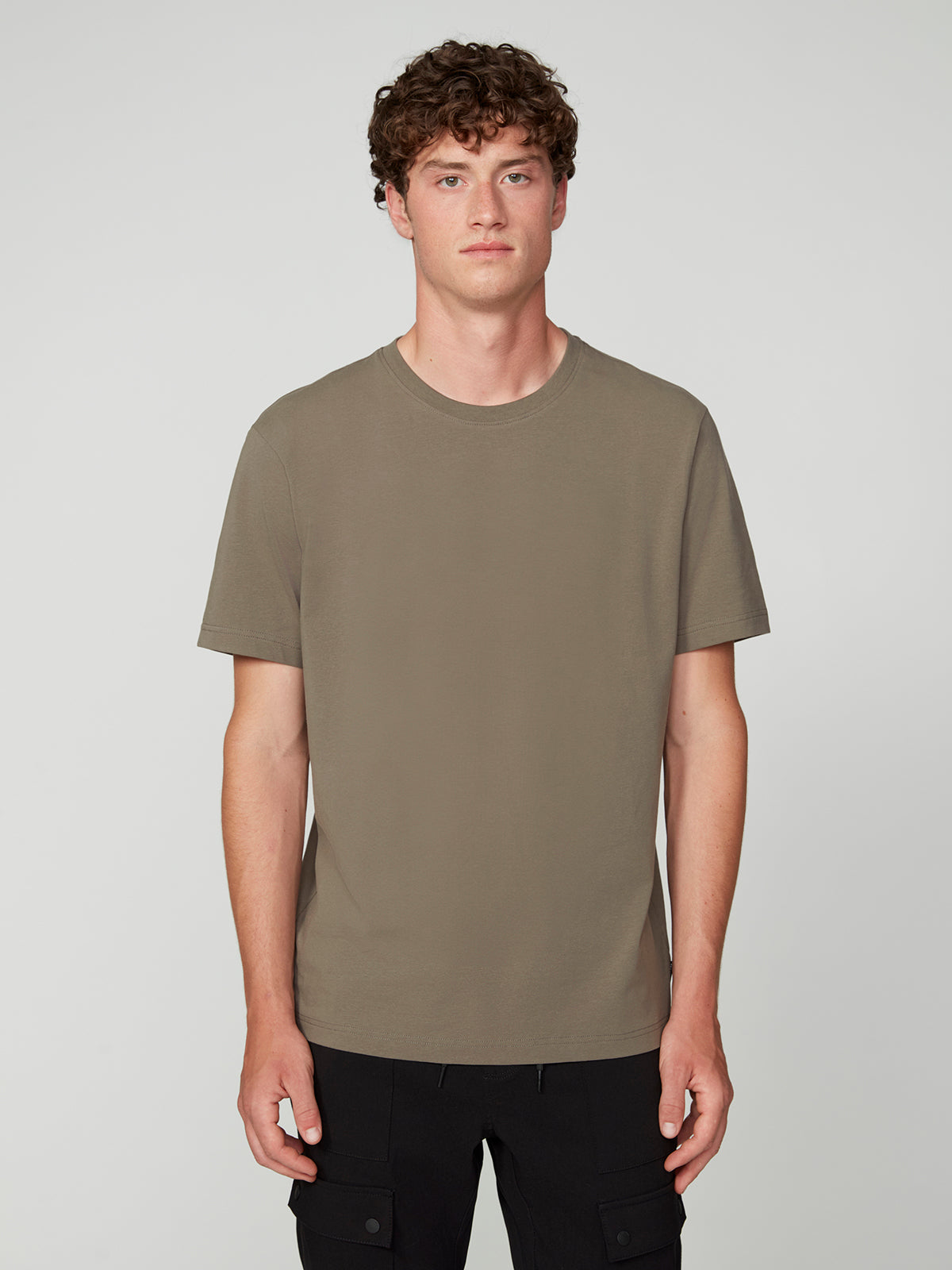 A young man with curly brown hair wears a PROJEK RAW MENS SHORT SLEEVE CREW NECK T-SHIRT in olive green and black pants, standing facing forward against a light gray background for a casual look.
