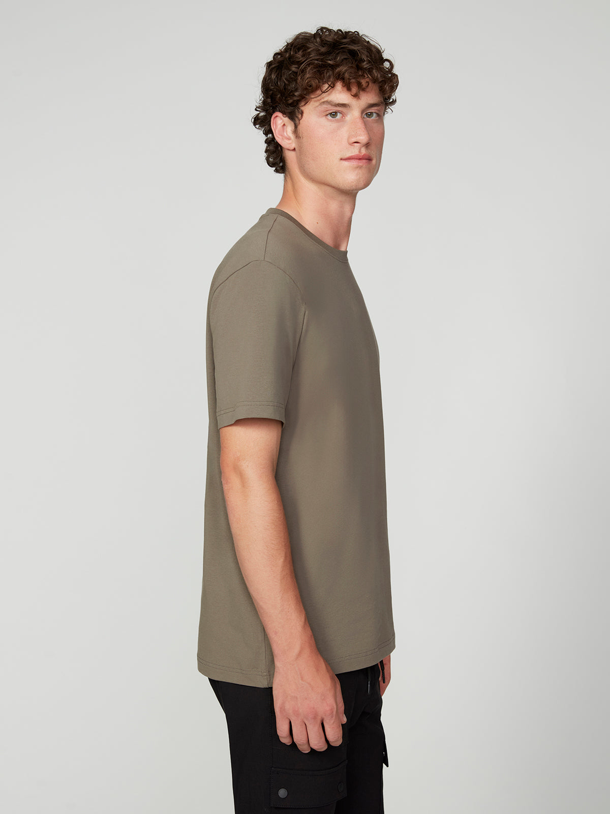 A young man with curly brown hair stands in side profile, wearing the PROJEK RAW MENS SHORT SLEEVE CREW NECK T-SHIRT in taupe and black pants against a light gray background—an effortless staple for any casual wardrobe.