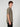 A young man with curly brown hair stands in side profile, wearing the PROJEK RAW MENS SHORT SLEEVE CREW NECK T-SHIRT in taupe and black pants against a light gray background—an effortless staple for any casual wardrobe.