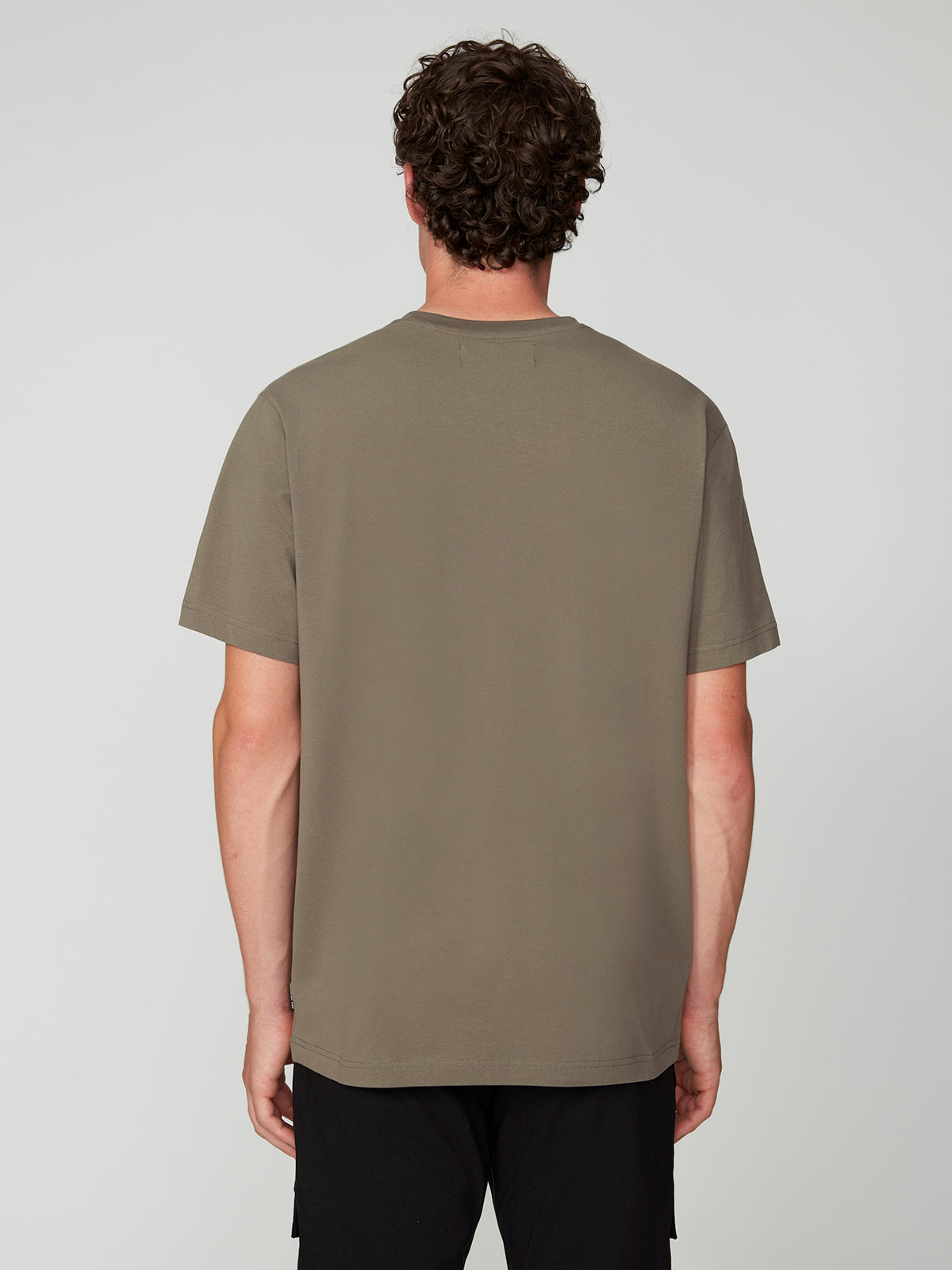 A person with short curly hair faces away from the camera, wearing a PROJEK RAW MENS SHORT SLEEVE CREW NECK T-SHIRT in olive green and black pants against a light gray background—perfect for any casual wardrobe.