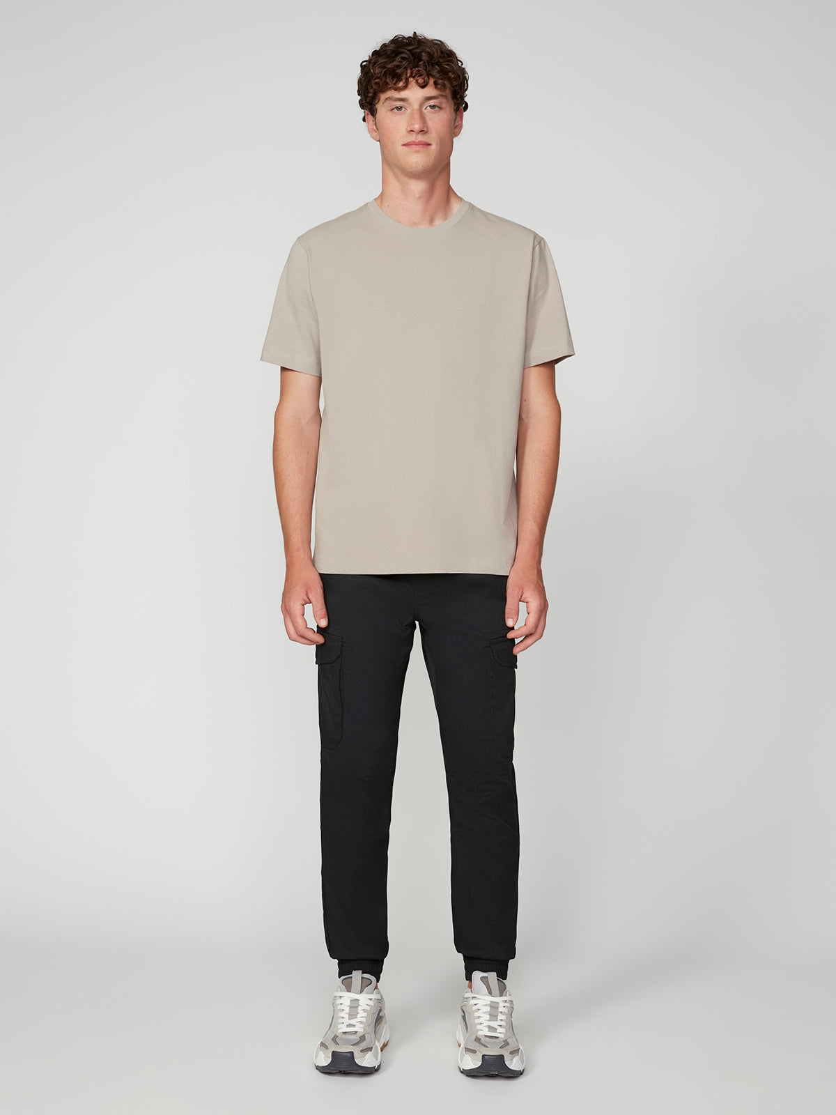 A young man stands facing forward against a light gray background, wearing the PROJEK RAW MENS SHORT SLEEVE CREW NECK T-SHIRT in beige, paired with black pants and white sneakers—ideal for any casual wardrobe.
