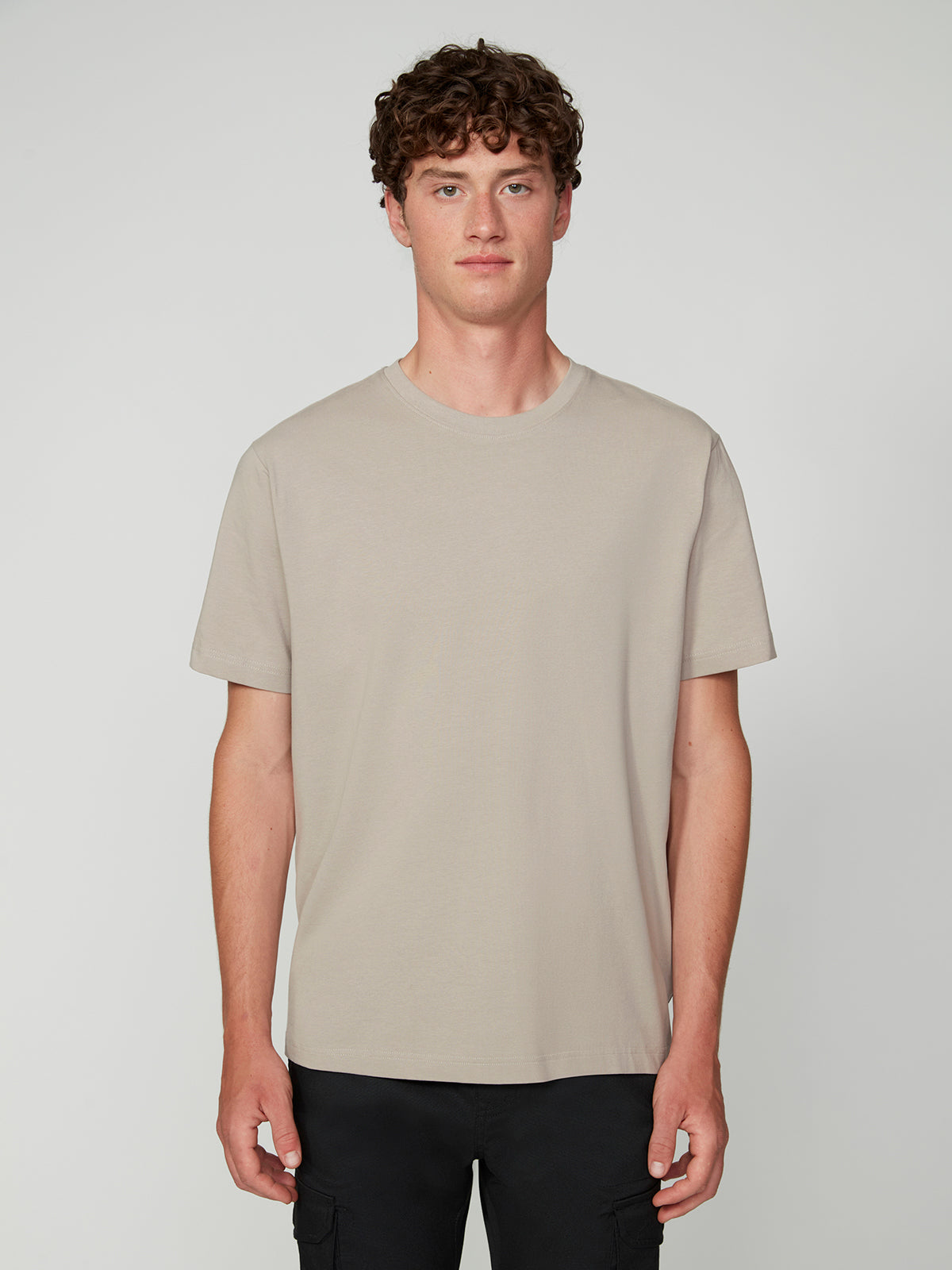 A young man with curly brown hair wears the PROJEK RAW MENS SHORT SLEEVE CREW NECK T-SHIRT in beige and black pants, standing against a light gray background—a versatile casual staple.